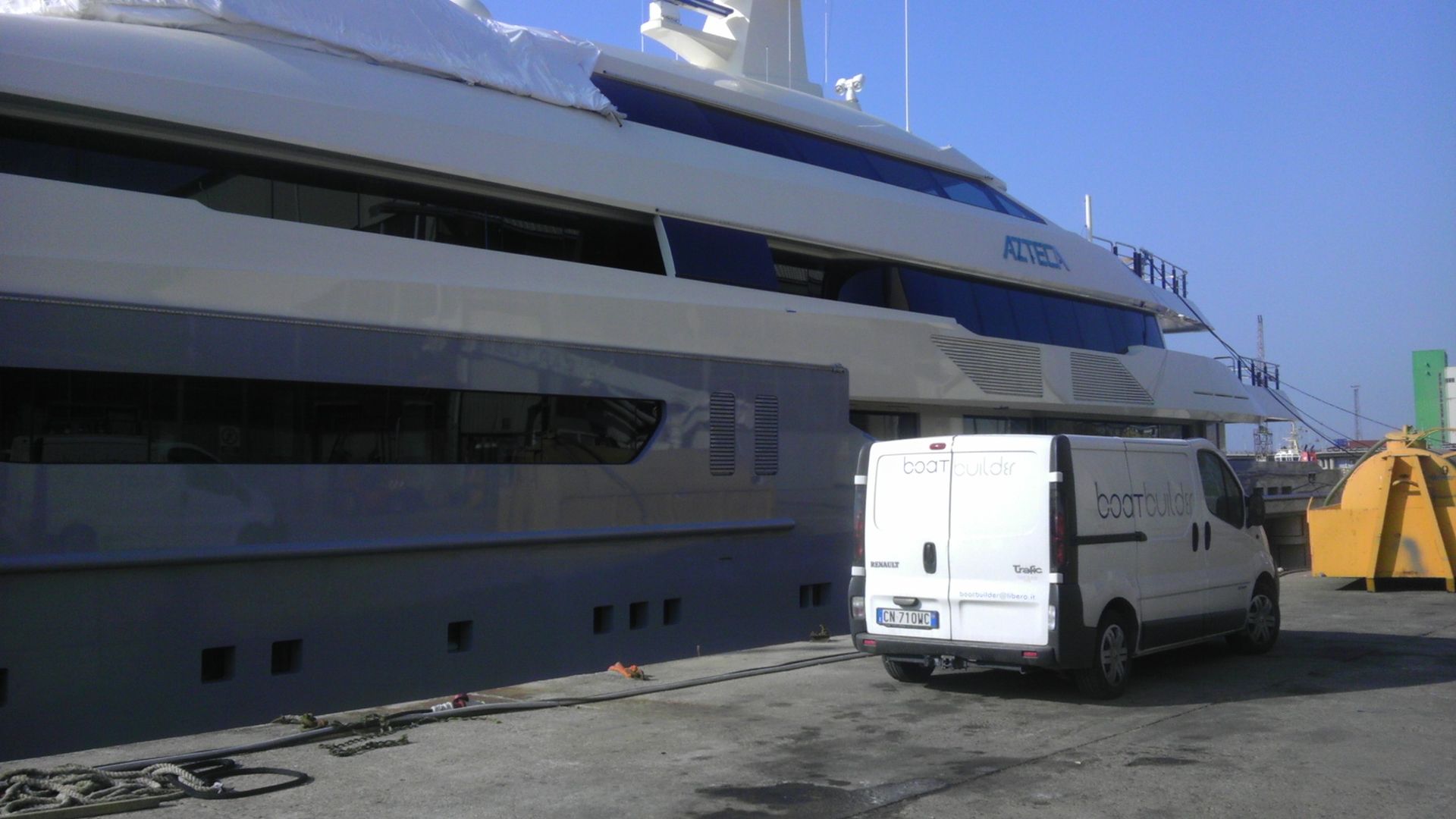 Un furgone bianco parcheggiato accanto a un grande yacht di lusso su un molo in una giornata di sole.