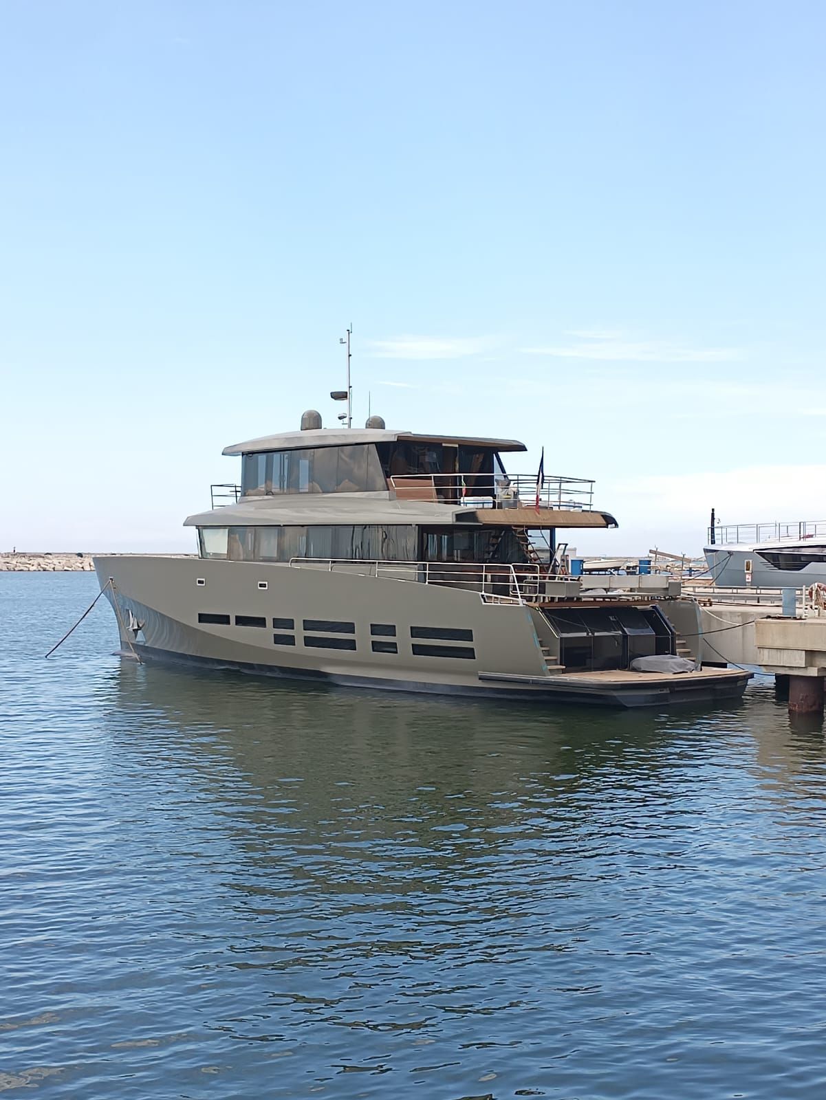 Un grande yacht grigio attraccato in acque calme; cielo azzurro e limpido.