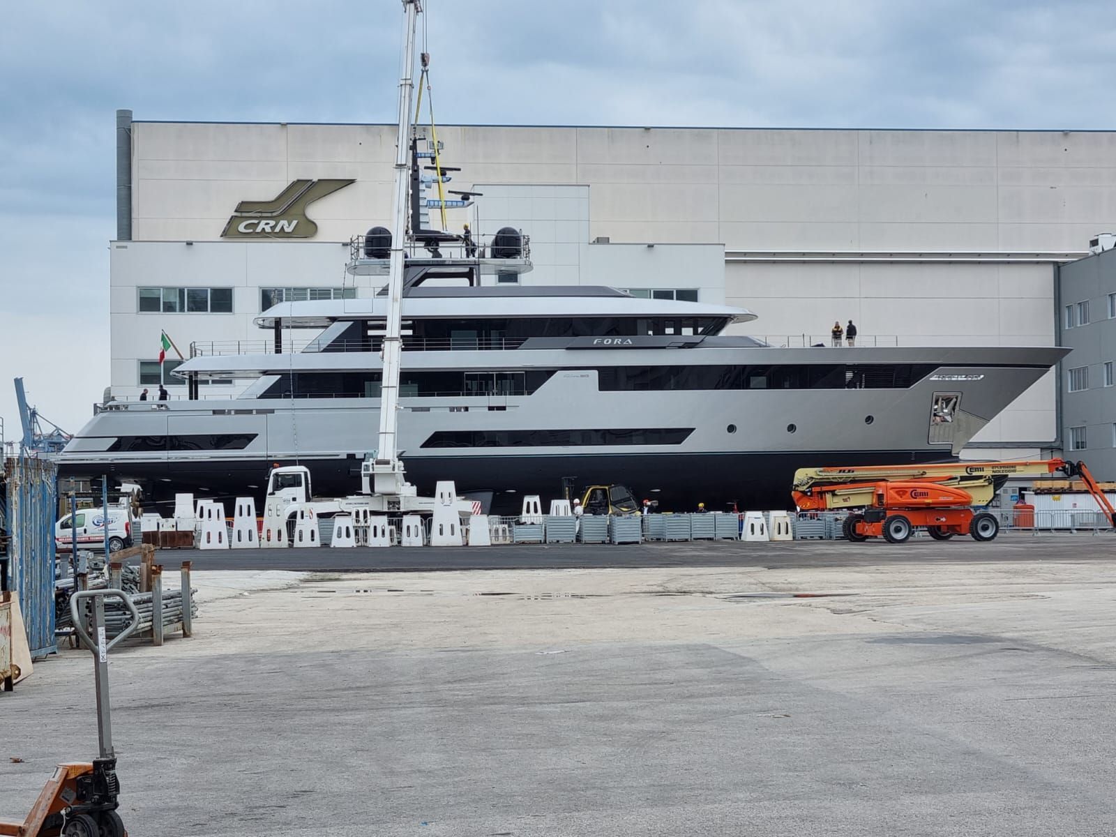 Grande yacht grigio su blocchi in un cantiere navale, con gru e attrezzature industriali.
