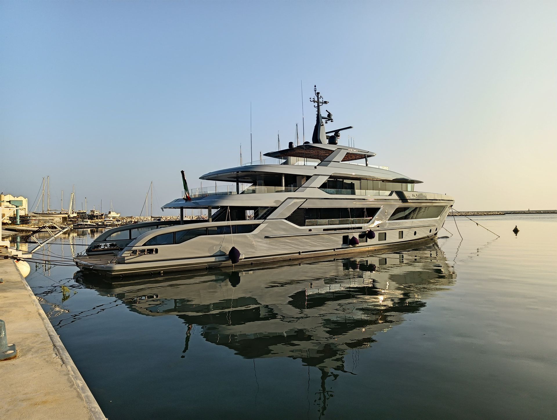 Un grande yacht grigio attraccato in acque calme, che ne riflette la forma; molo in primo piano, cielo limpido.