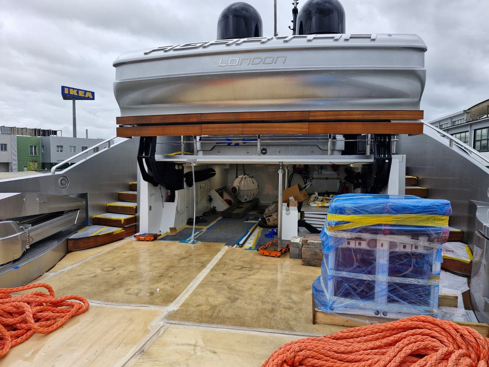 Vista posteriore di uno yacht, con tender, ponte di legno, corde arancioni e materiali da costruzione su un molo.