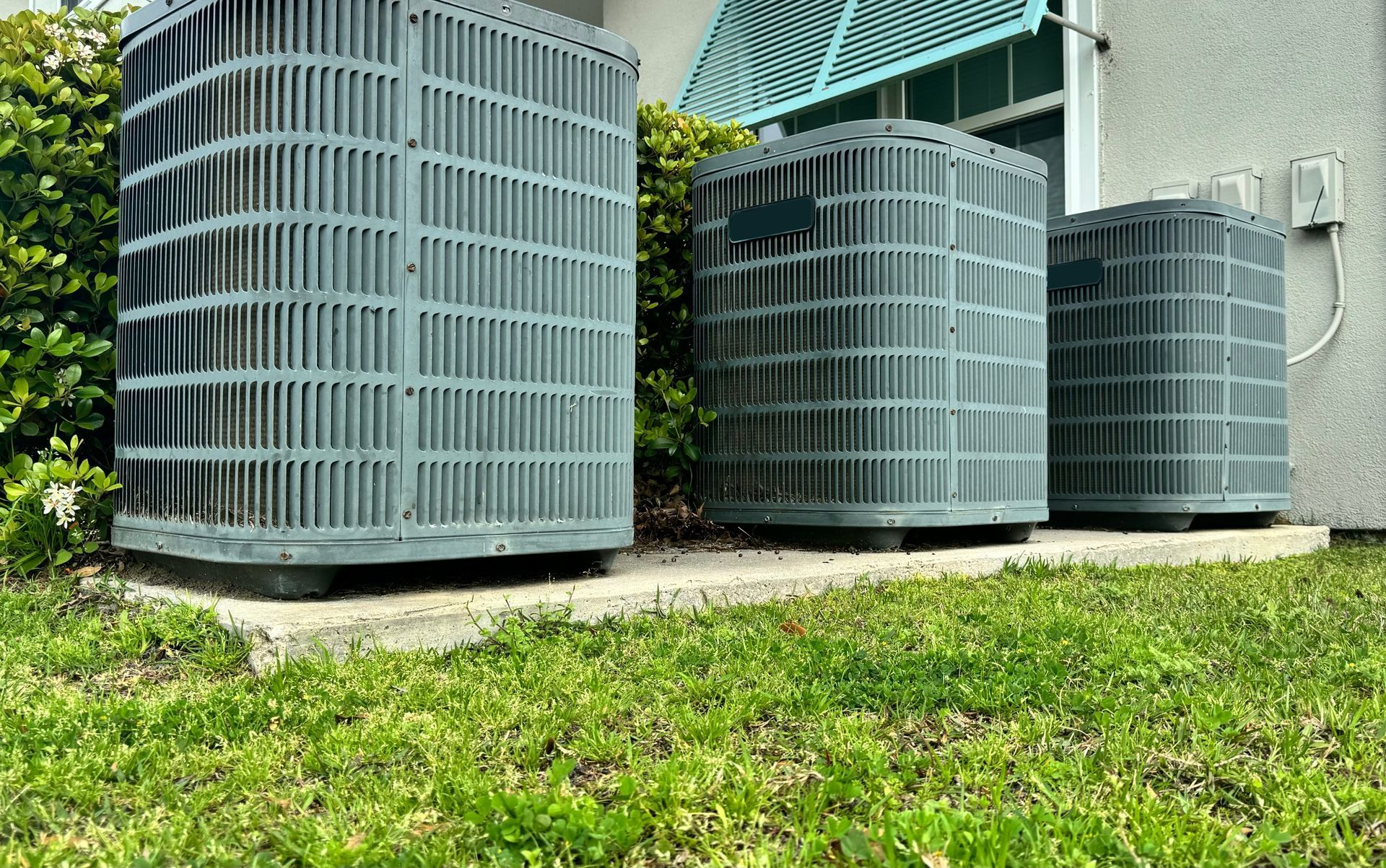 Three gray air conditioning units sitting on concrete in a grassy area next to a building.