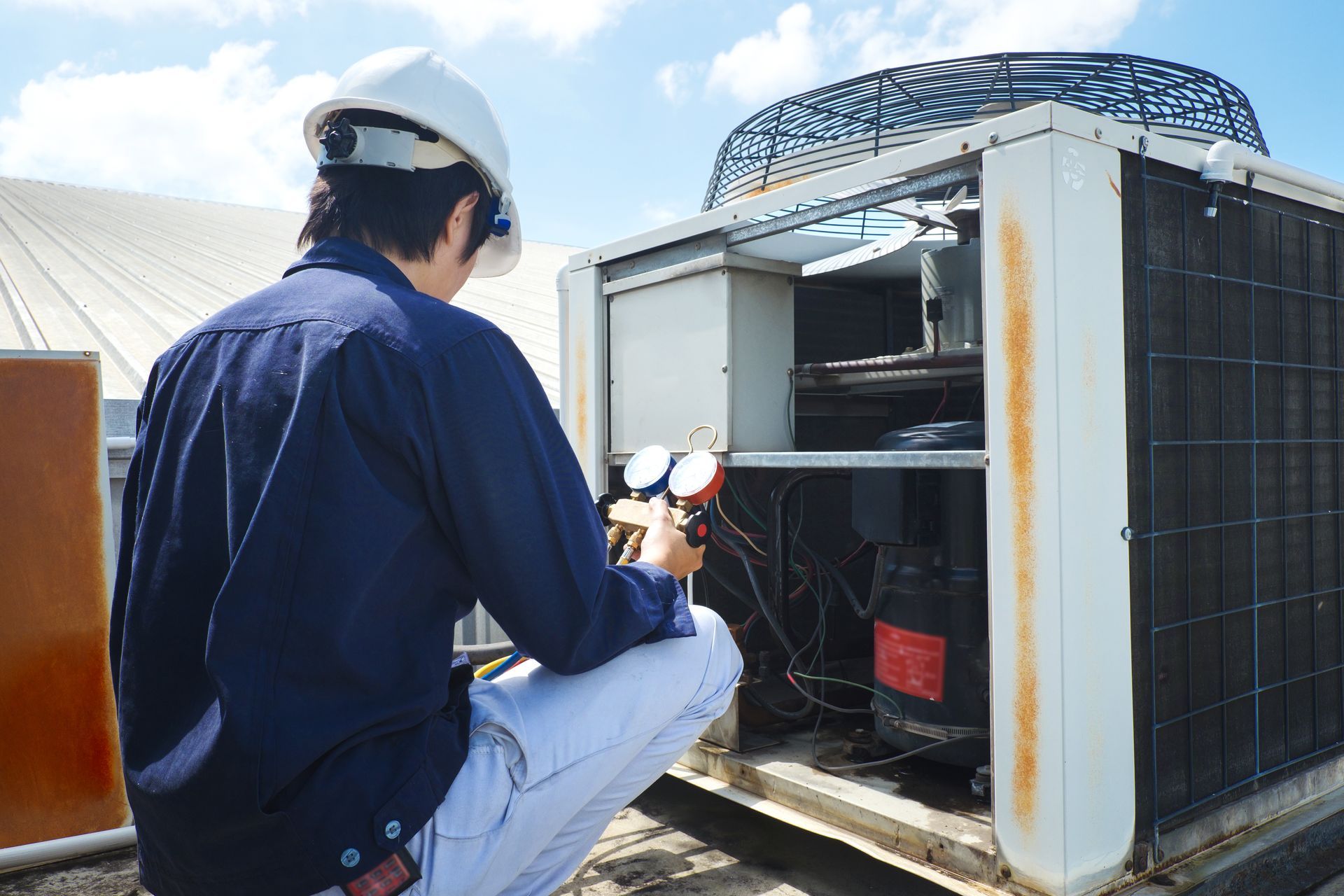 HVAC technician in a hard hat inspects rooftop air conditioning unit with gauges. Sunny day.