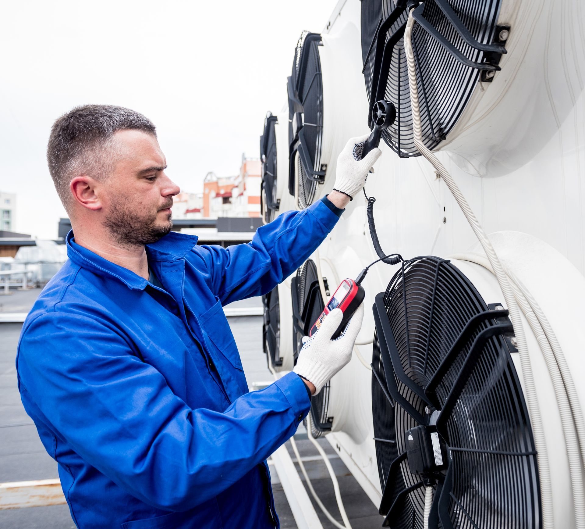 Man in blue jumpsuit inspects HVAC unit, using a meter and wearing gloves on a rooftop.