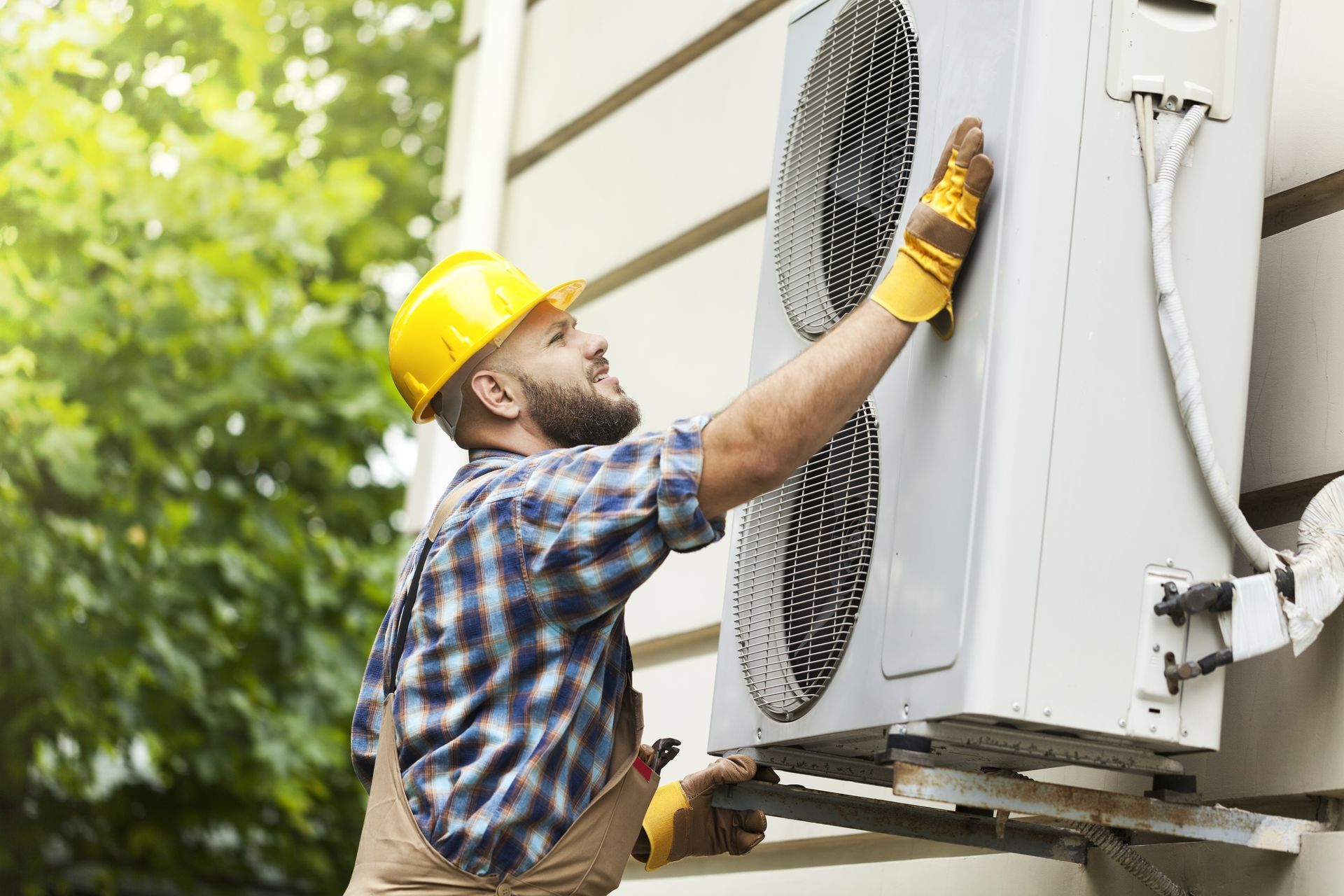 HVAC technician in yellow hard hat inspecting an outdoor air conditioning unit.