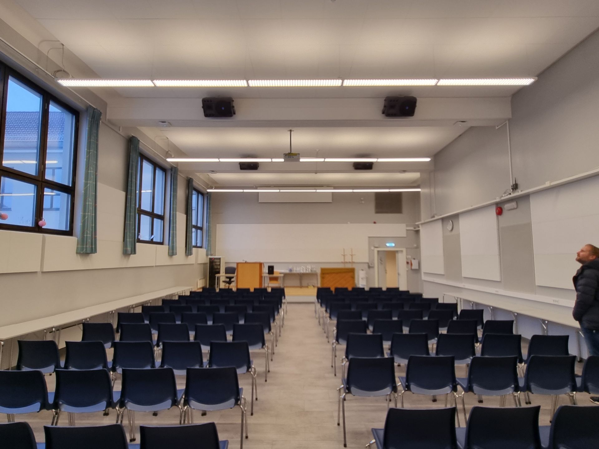 Auditorium med rader med mørkeblå stoler, vinduer og en person som står nær høyre vegg.