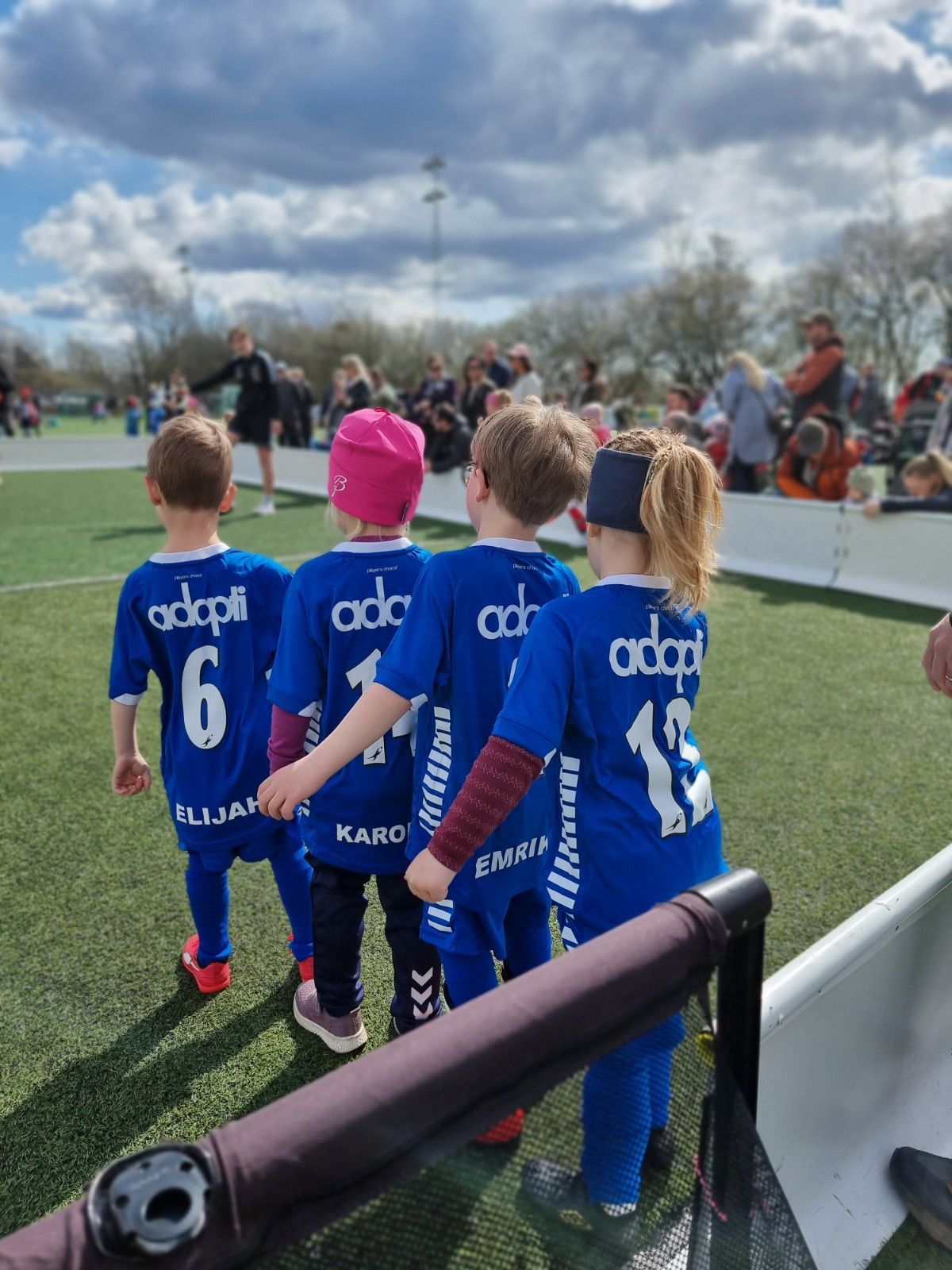Fire små barn i blå fotballdrakter går på en grønn bane. Draktene har «Adopt» på ryggen.
