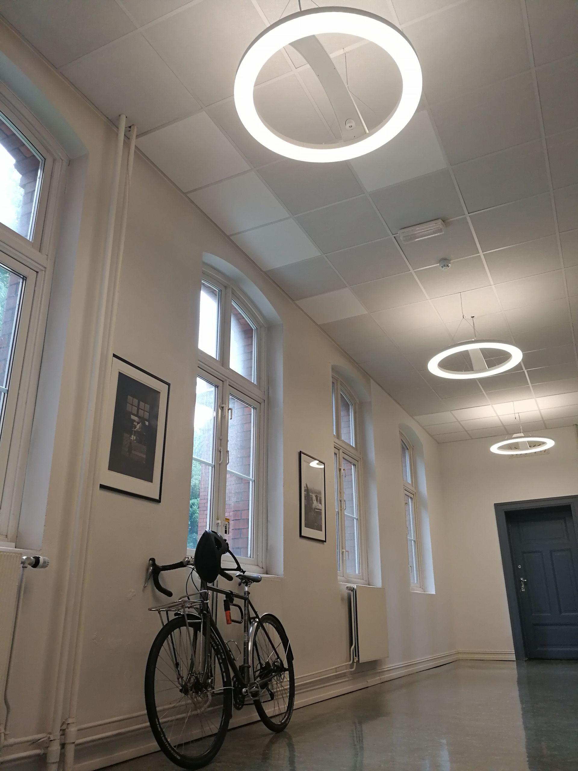 Gang med hvite vegger, vinduer og sirkulære lys; en sykkel står parkert inntil veggen.