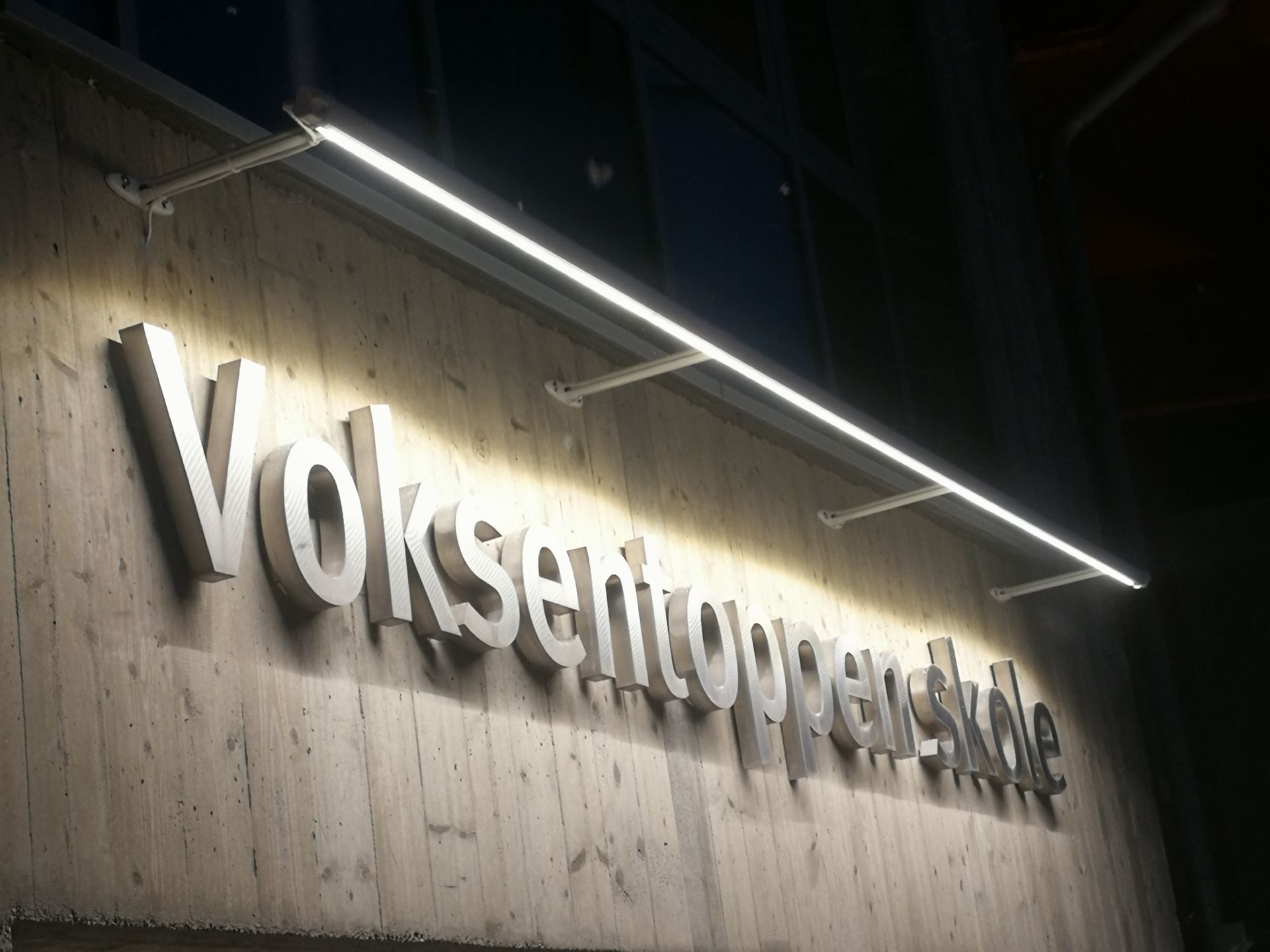 Opplyst skilt «Voksentoppen» på en betongvegg, med en lysstripe over.
