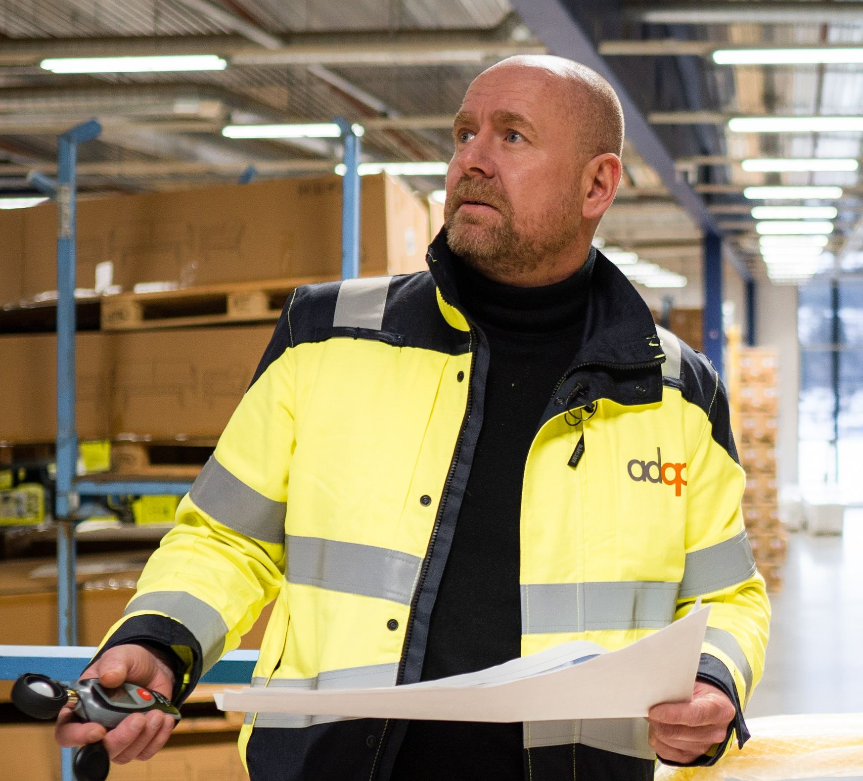 Mann i gul refleksjakke som holder papirer, ser opp i et lagerlokale.