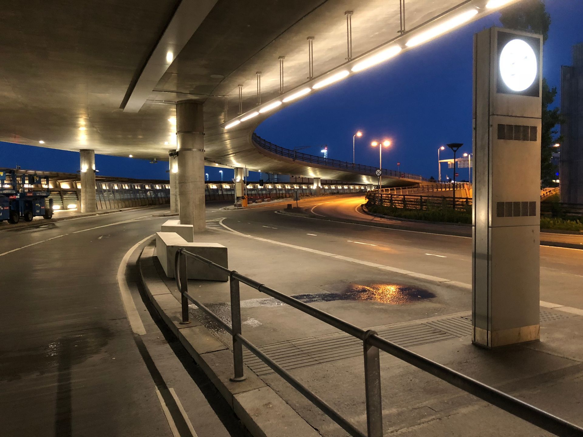 Nattbilde av en avleveringssone på flyplassen. Betongsøyler og opplyst takkonstruksjon. Veien er våt.