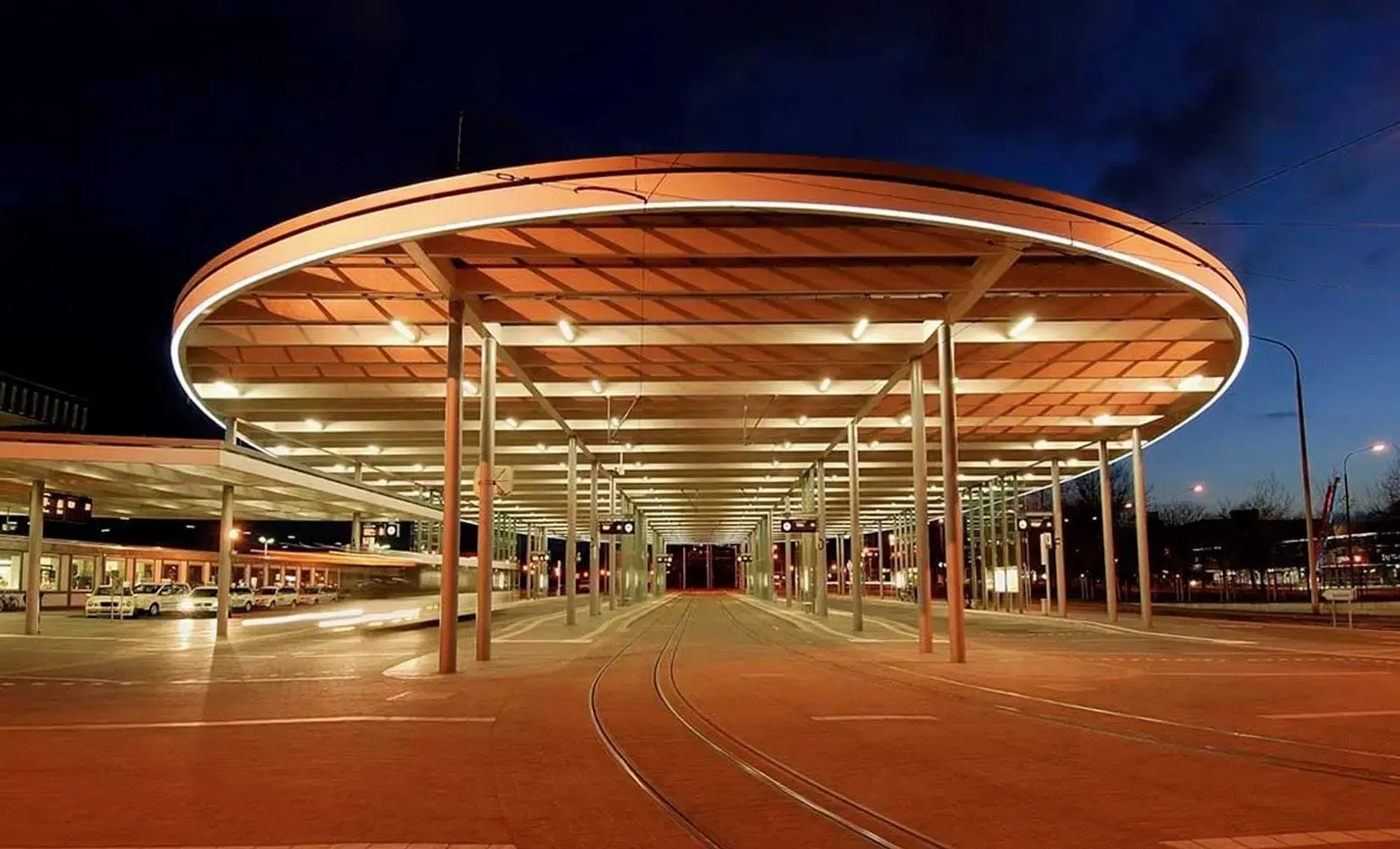 Moderne busstasjon opplyst i skumringen; en stor, sirkulær baldakin støttet av søyler.