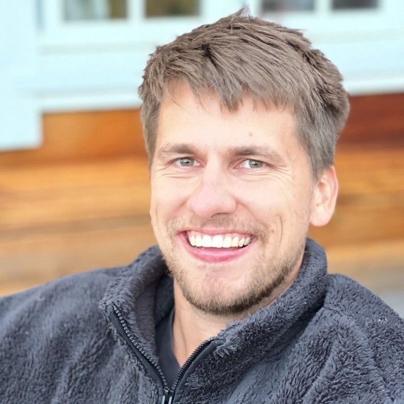 Mann med et varmt smil, iført en grå fleecejakke, utendørs med uskarp bakgrunn.