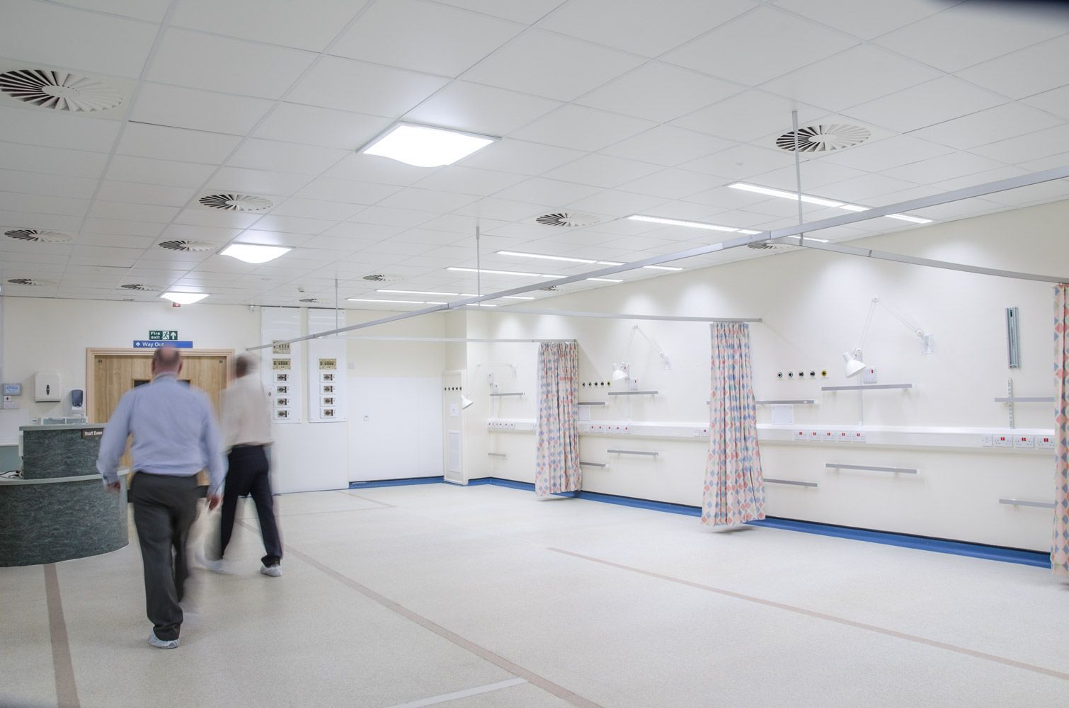 To personer går gjennom et lyst sykehusrom med medisinsk utstyr og pasientskillevegger.