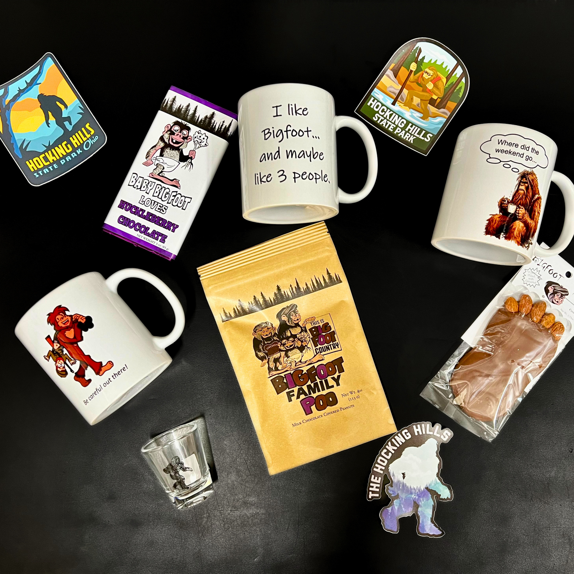 Bigfoot-themed merchandise on a black surface includes mugs, stickers, chocolate, and a shot glass.