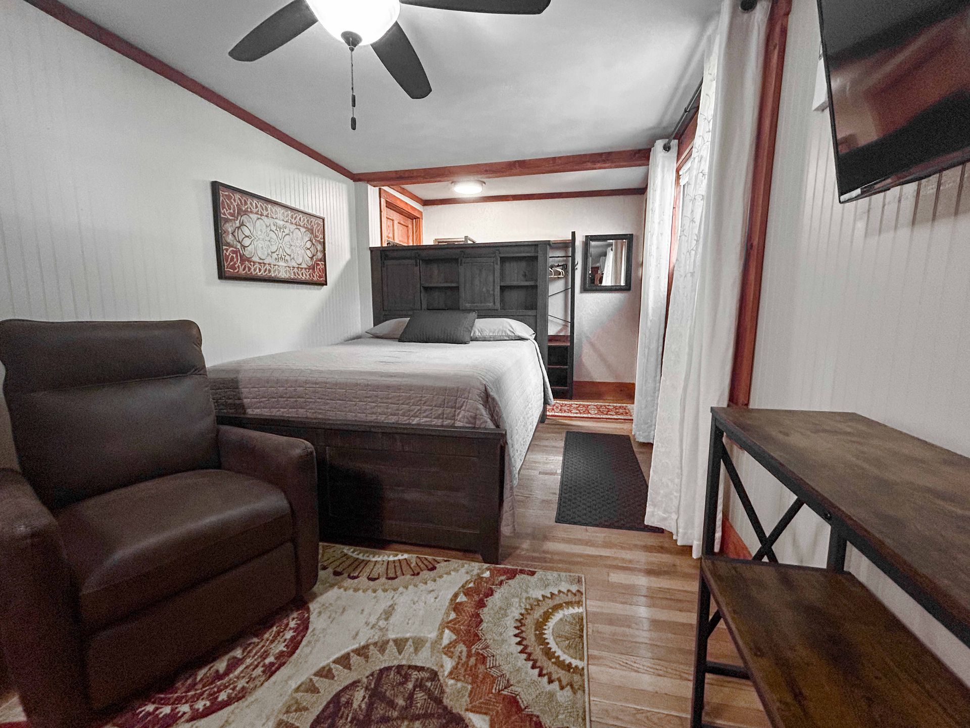 A small bedroom with a brown recliner, bed, and wooden bench. The walls are white with red trim.