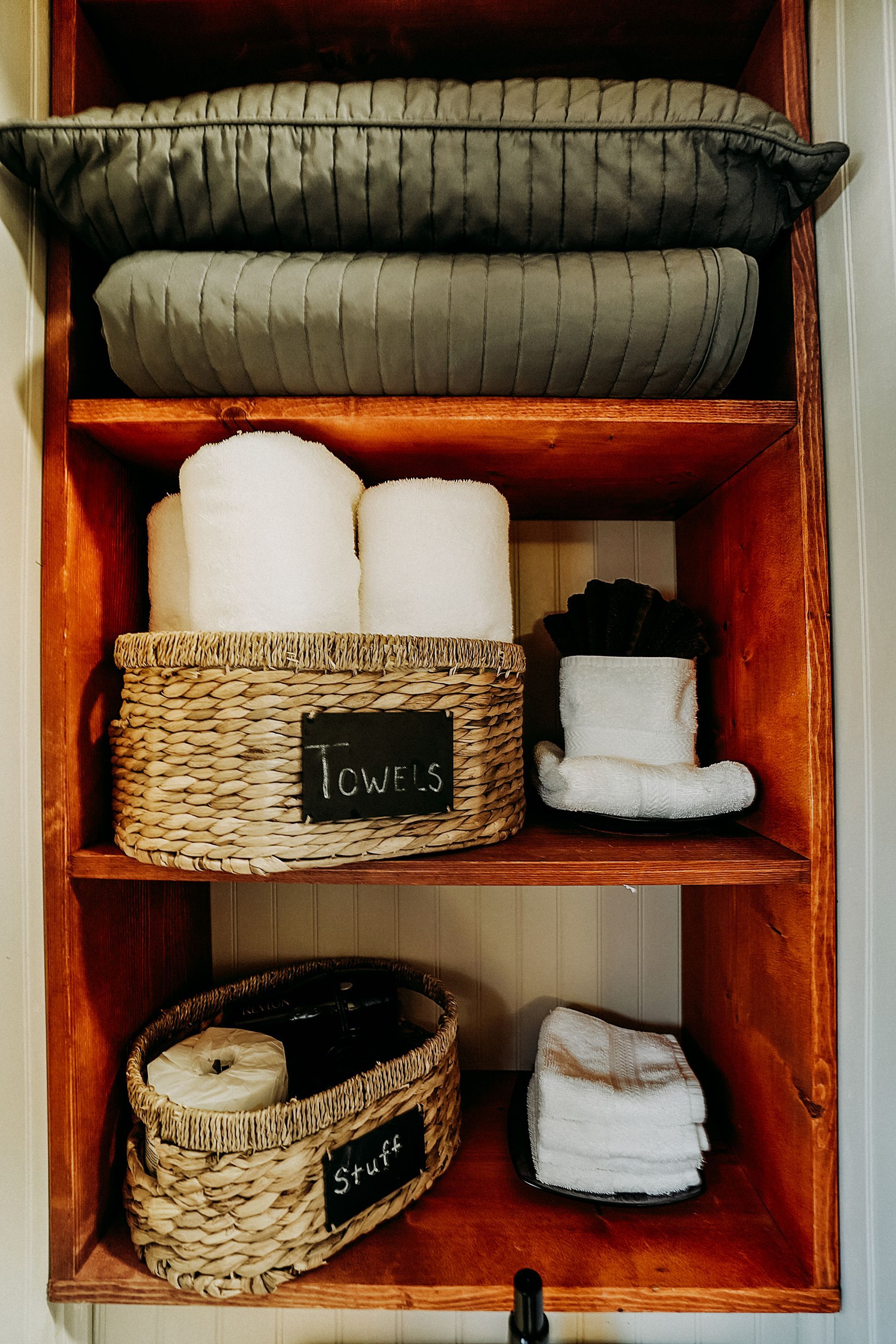 Wooden bathroom shelf with towels, toilet paper, and decorative pillows. Warm-toned wood with white and gray textiles.
