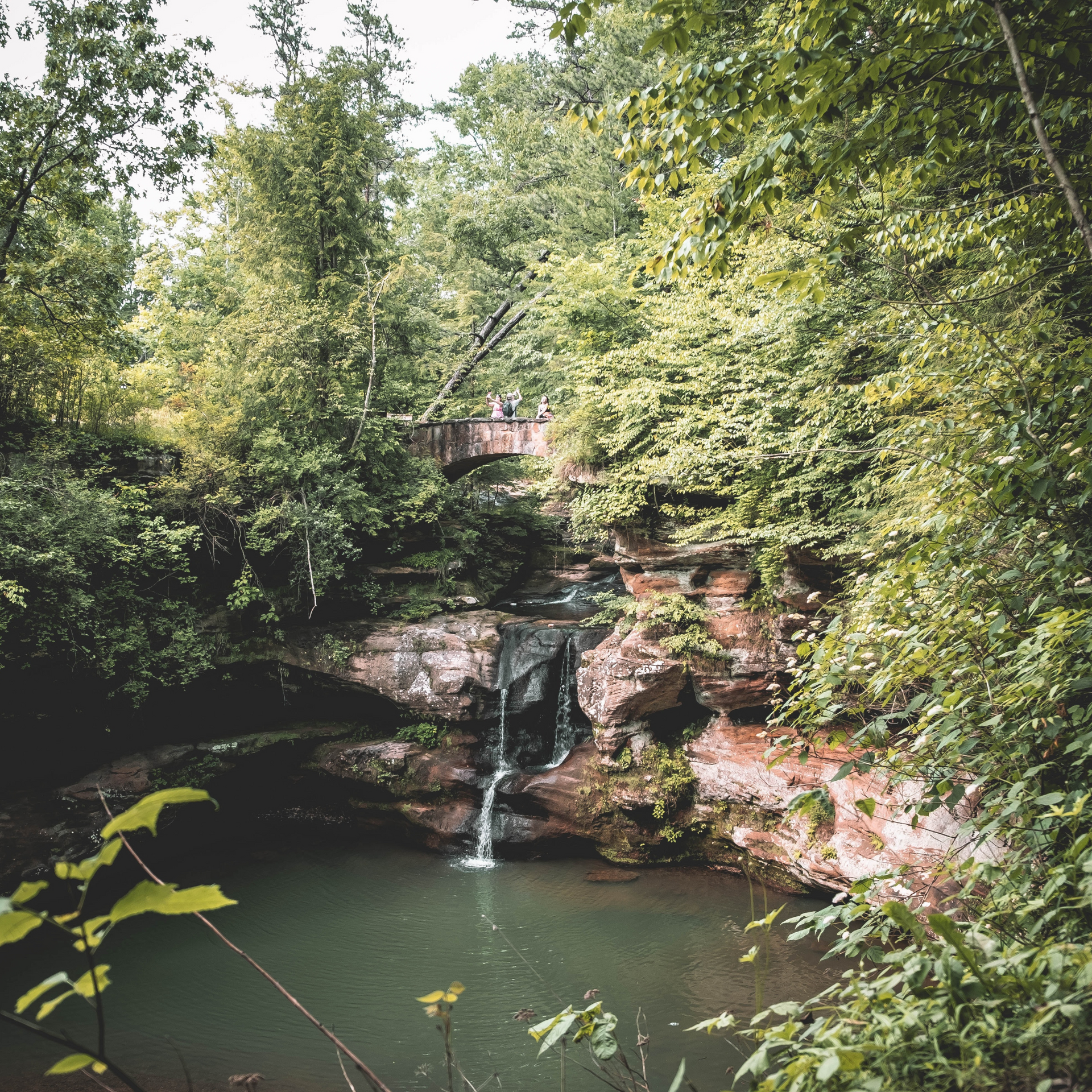 Waterfall cascading into a small pool surrounded by lush green trees. A stone bridge is visible in the background.