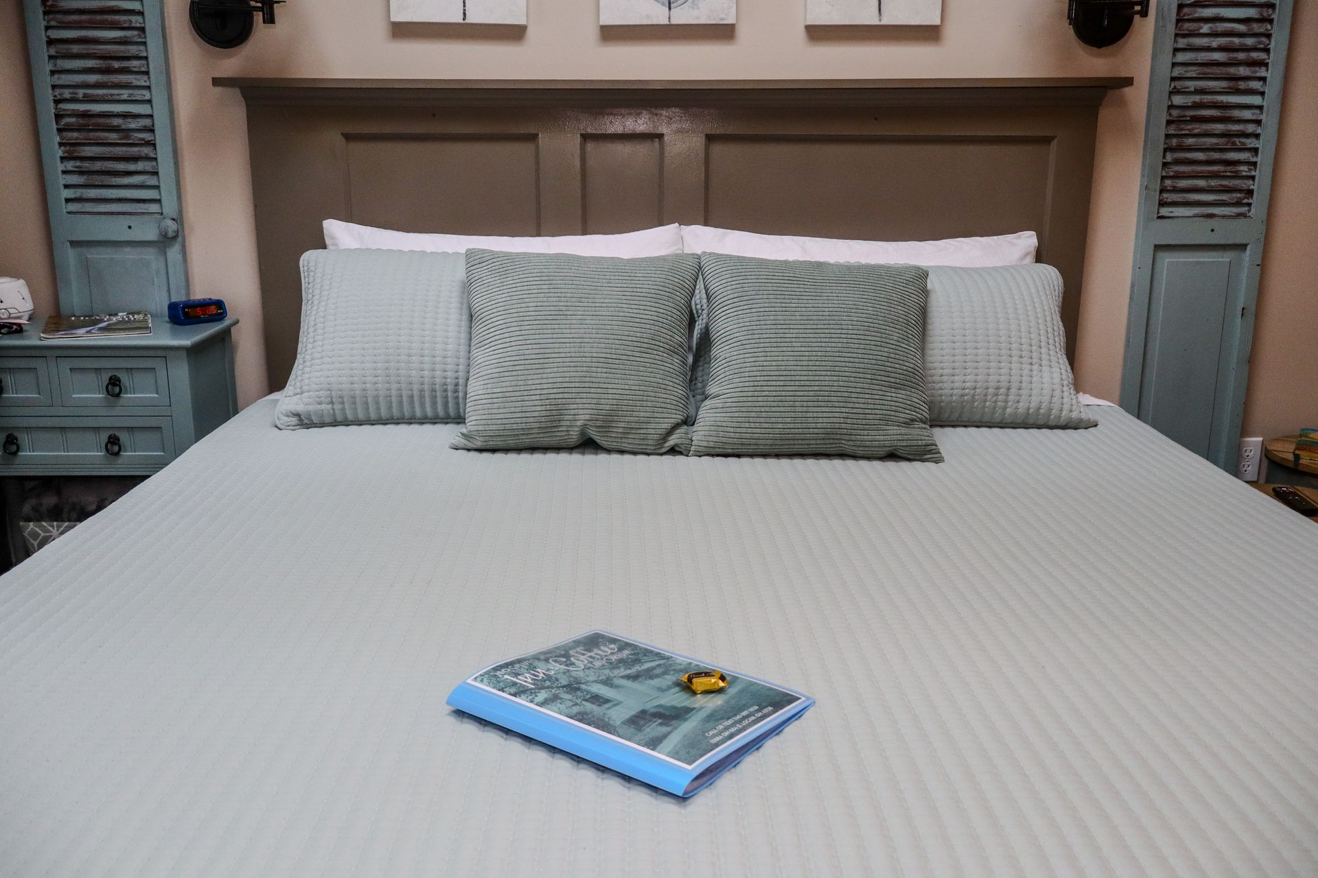 A bed with a book and pillows, set against a headboard and shutters. The bedding is light green and the book is blue.