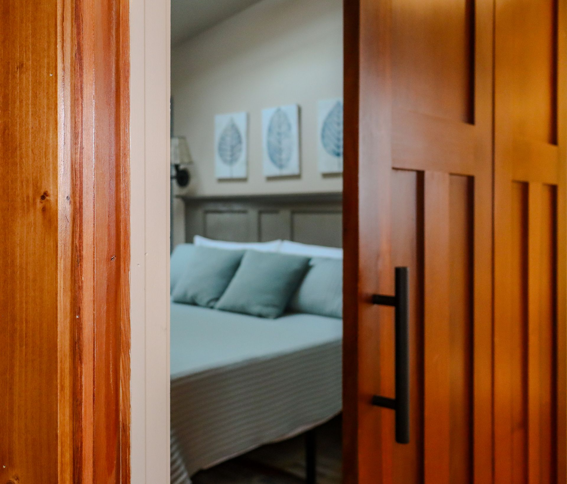 Open wooden door revealing a bedroom with a bed, pillows, and artwork; warm lighting.