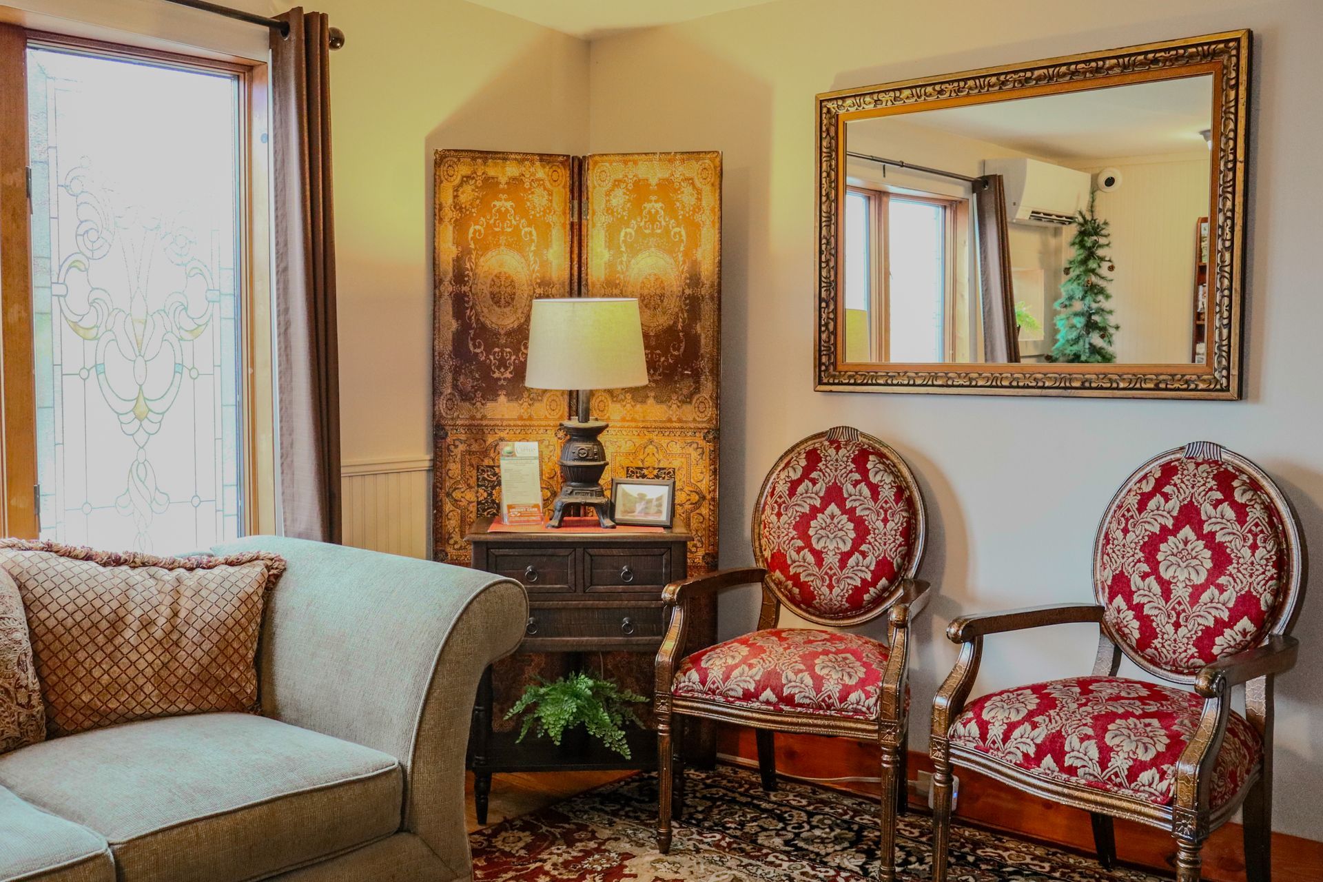 Cozy living room with a sofa, two ornate chairs, antique screen, and a mirror. The chairs have red patterned upholstery.