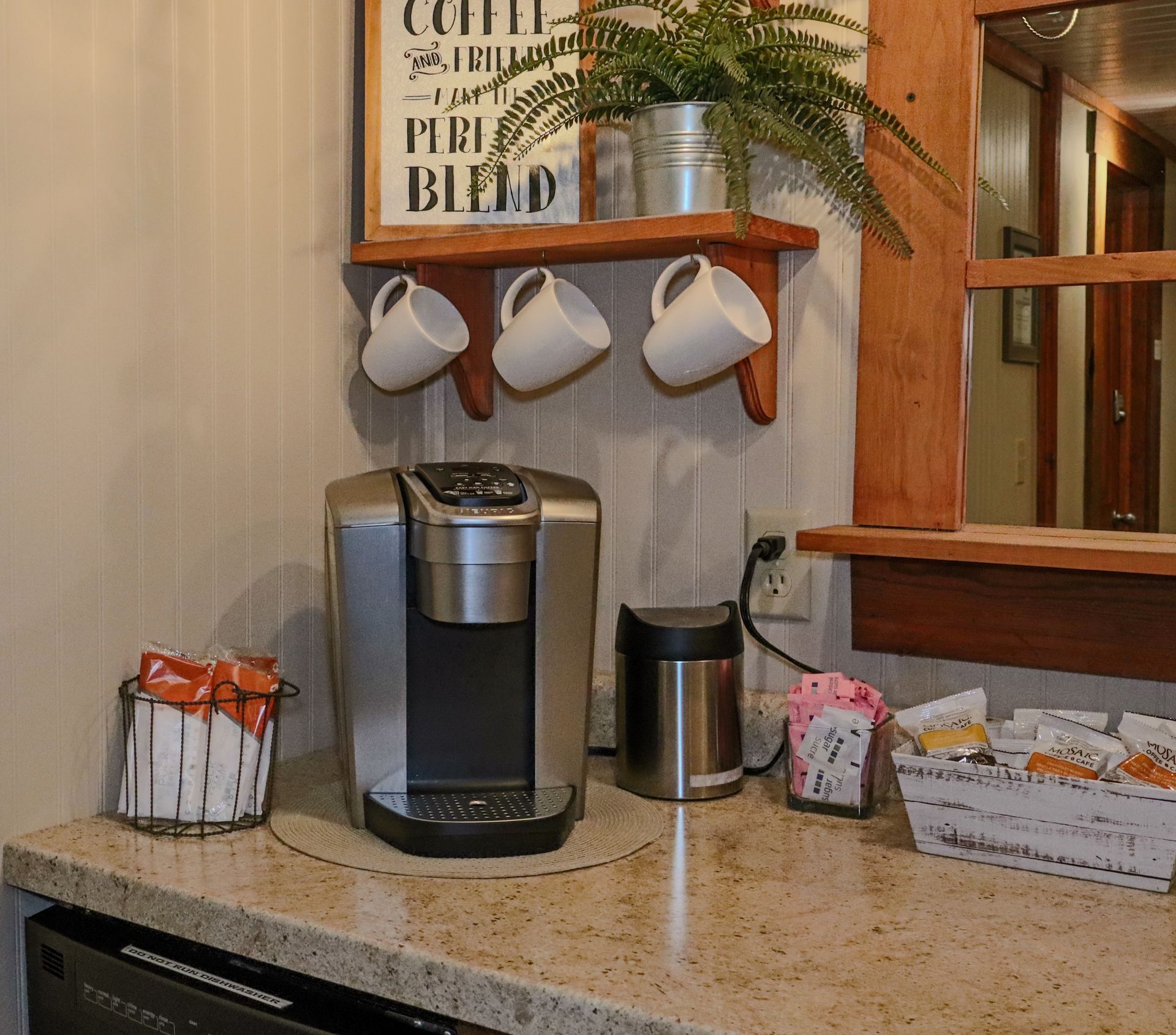 Coffee station with a Keurig, mugs, and coffee supplies on a countertop with a wooden shelf and window.