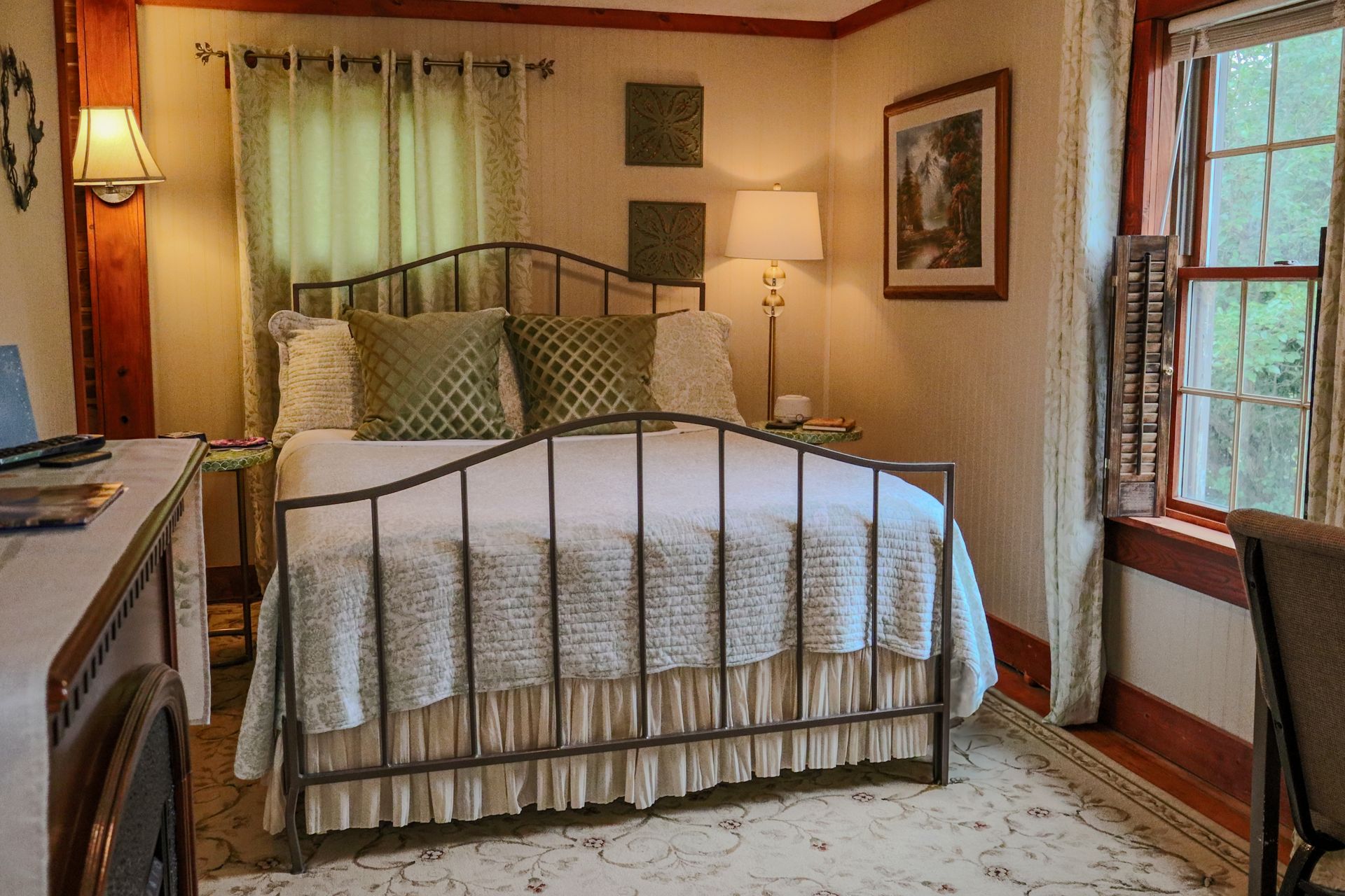 A bedroom with a bed, desk, and window. The bed has a metal frame, white bedding, and green pillows.