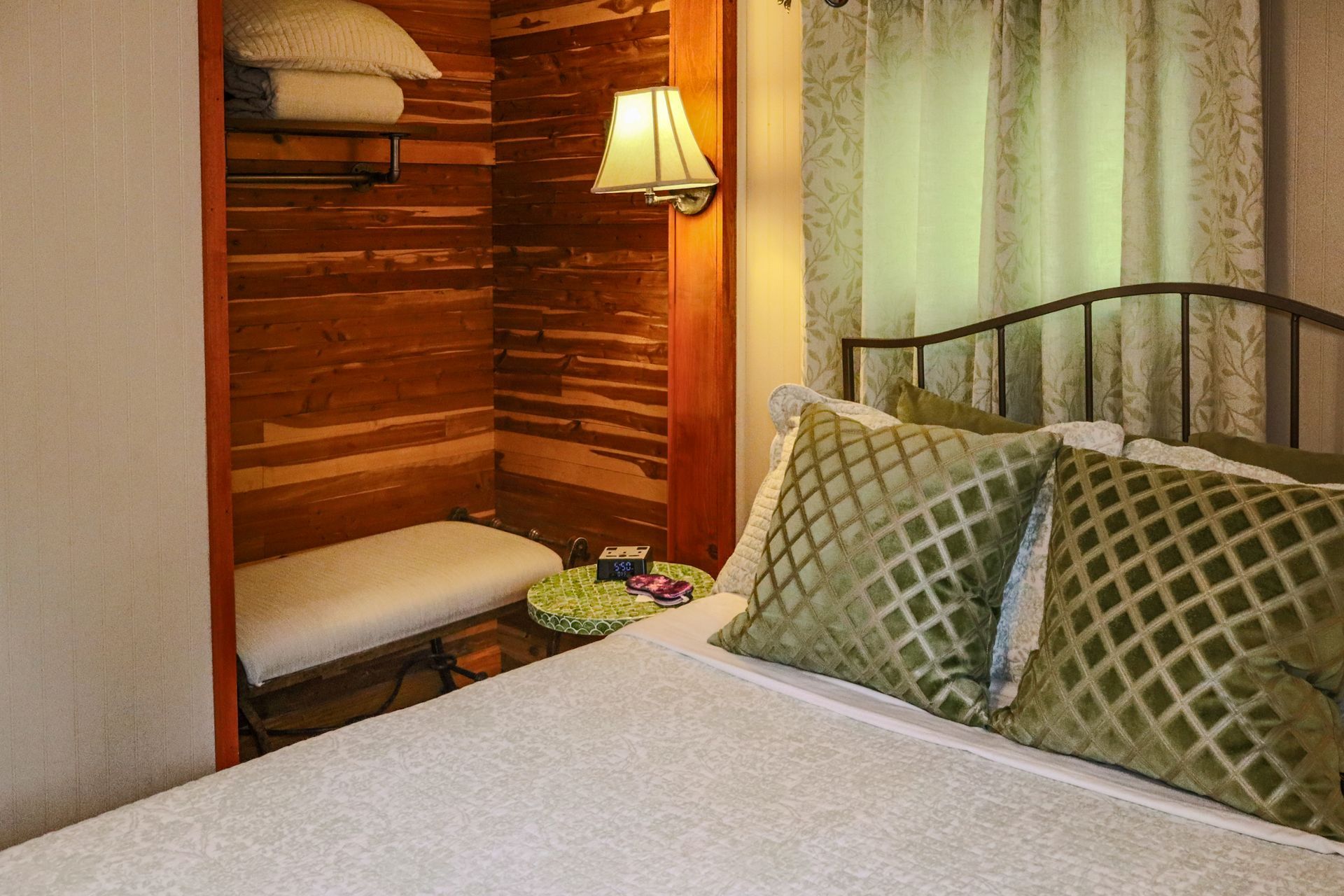 Bedroom with a bed, green pillows, and a built-in closet with a bench, towels, and a lamp. The closet has wooden paneling.