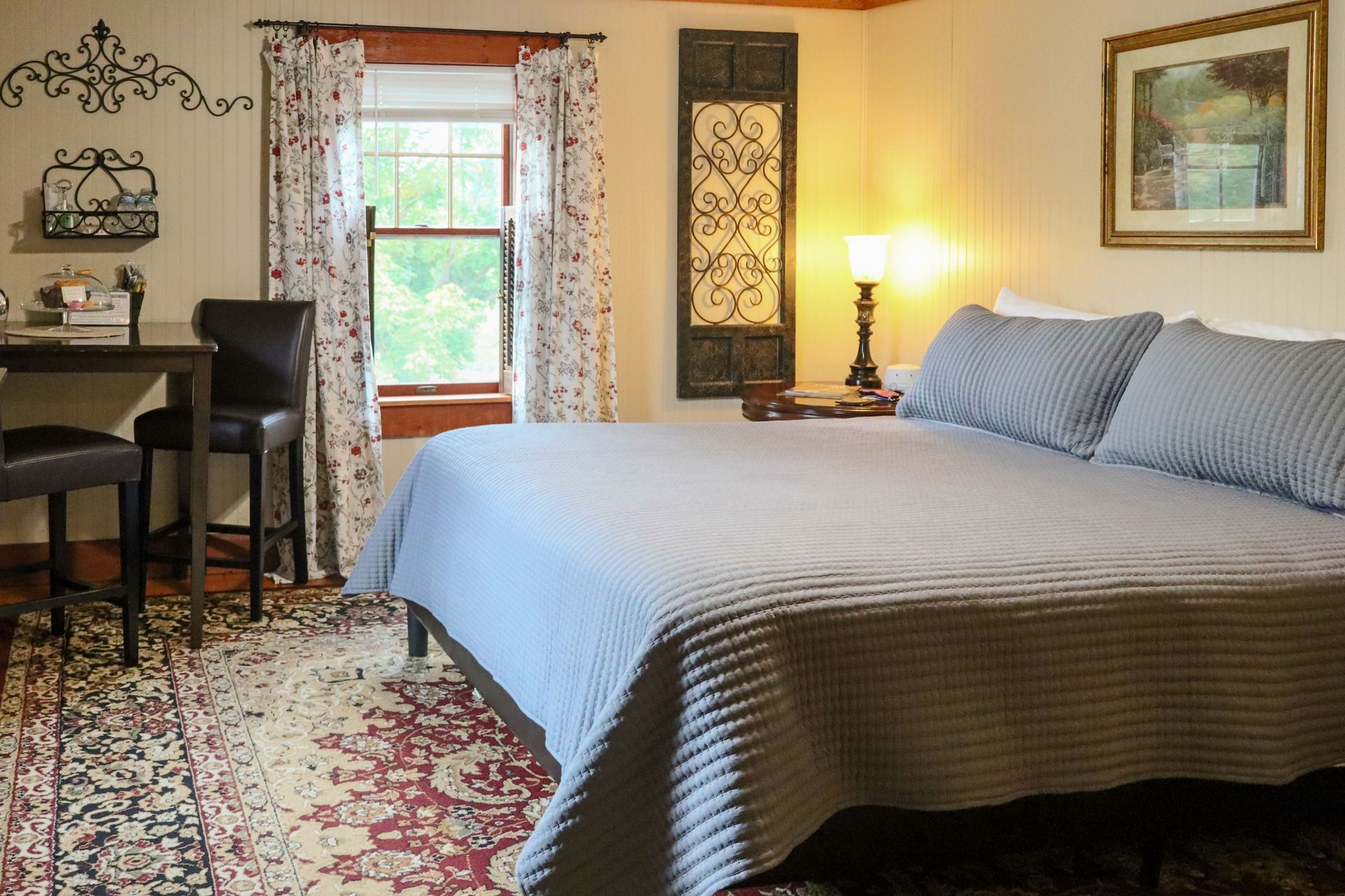 Cozy bedroom with king-sized bed, small table with chairs, window, and decorative rug. Soft lighting and floral curtains.