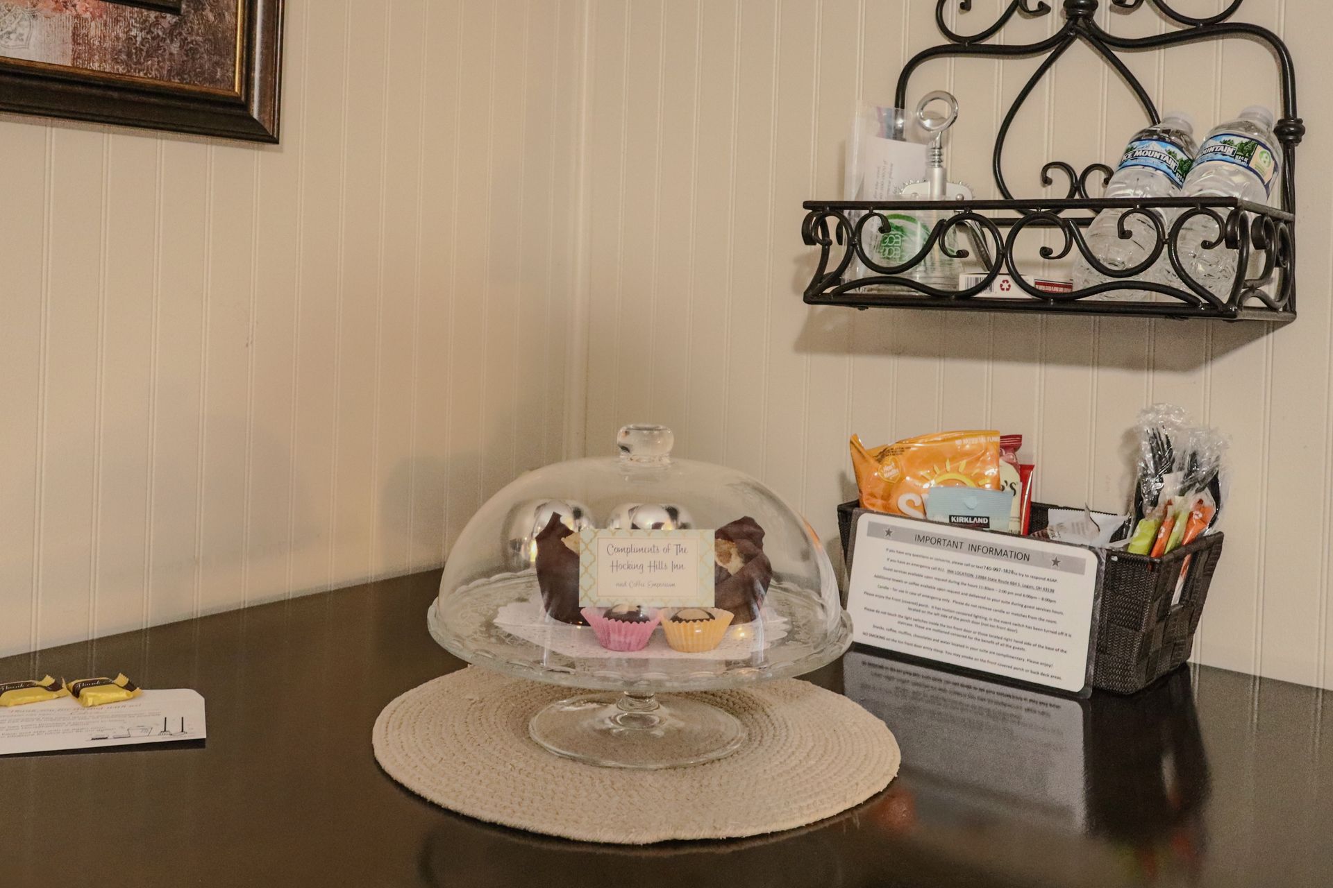 A cake stand with covered pastries on a table. A small shelf with glassware and snacks.