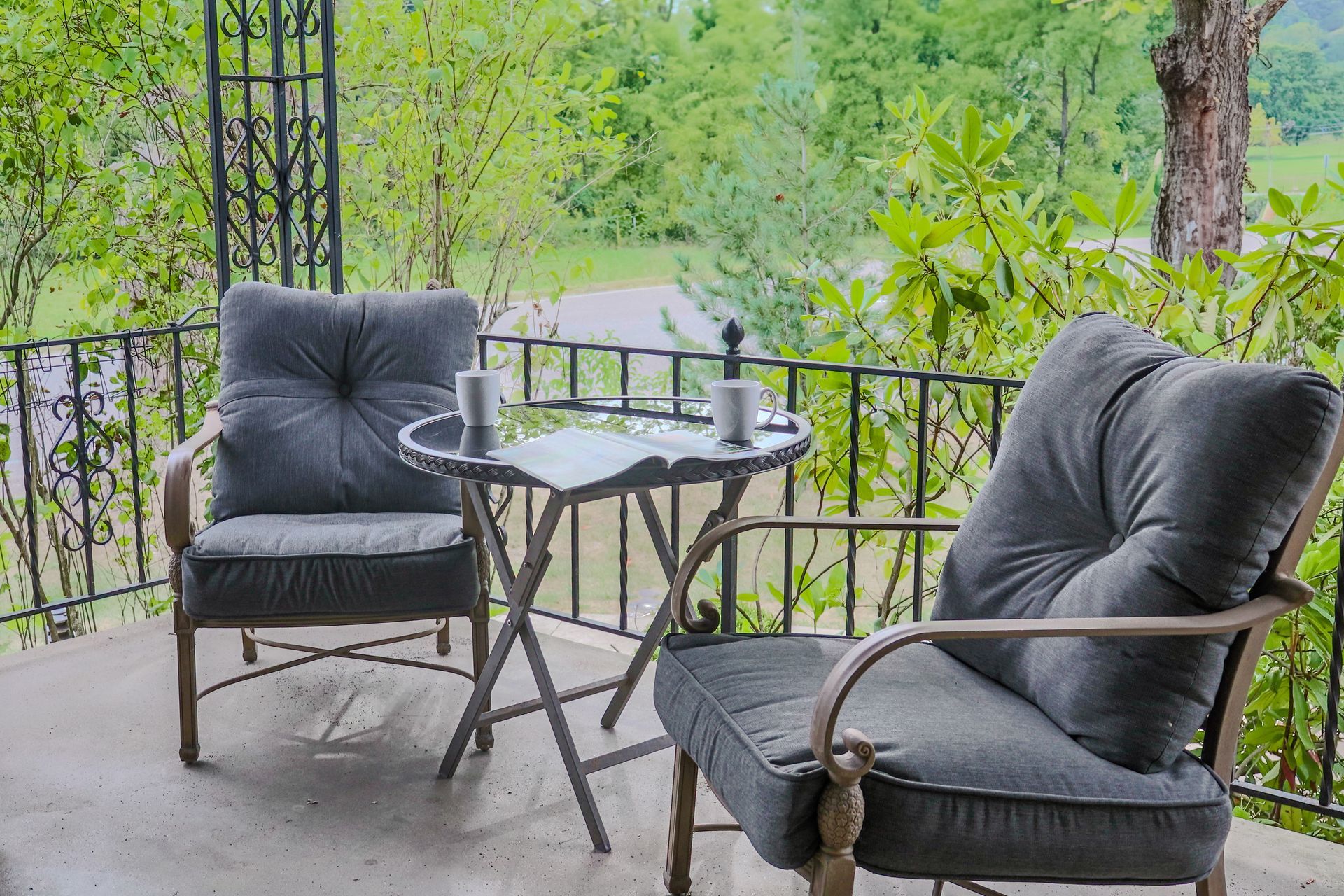 Two gray cushioned chairs and a small table sit on a porch, overlooking a green, leafy landscape. Two mugs sit on the table.