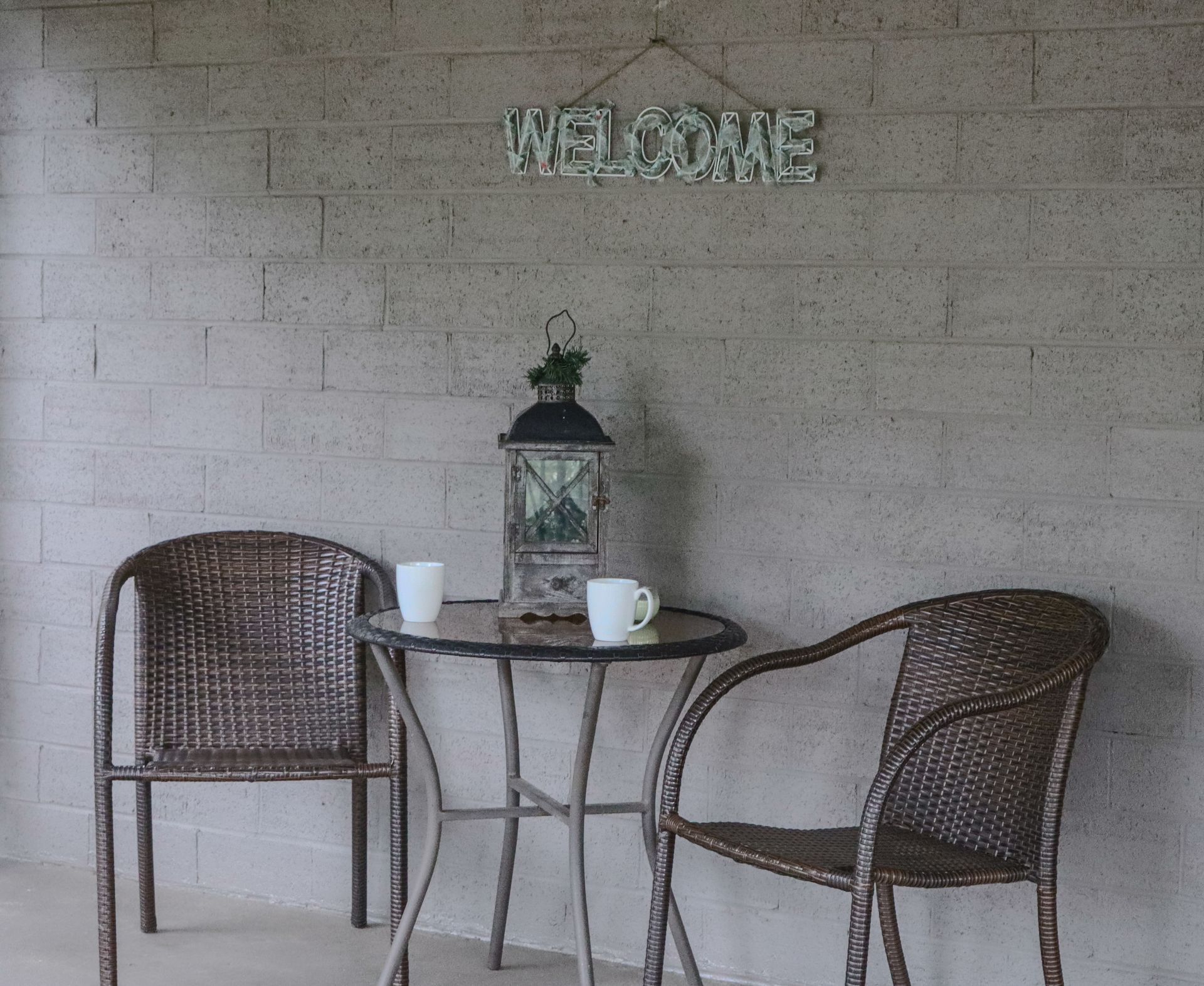 Two wicker chairs face a small table with a lantern and mugs. A welcome sign hangs above.
