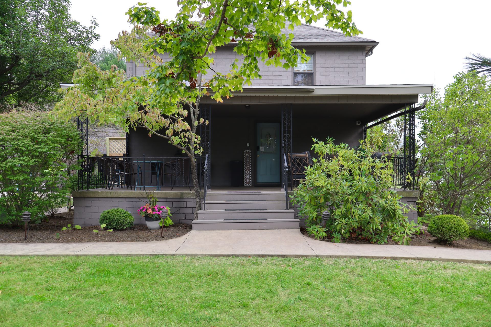 A two-story gray house with a porch, surrounded by green bushes and trees. A walkway leads to the front steps.