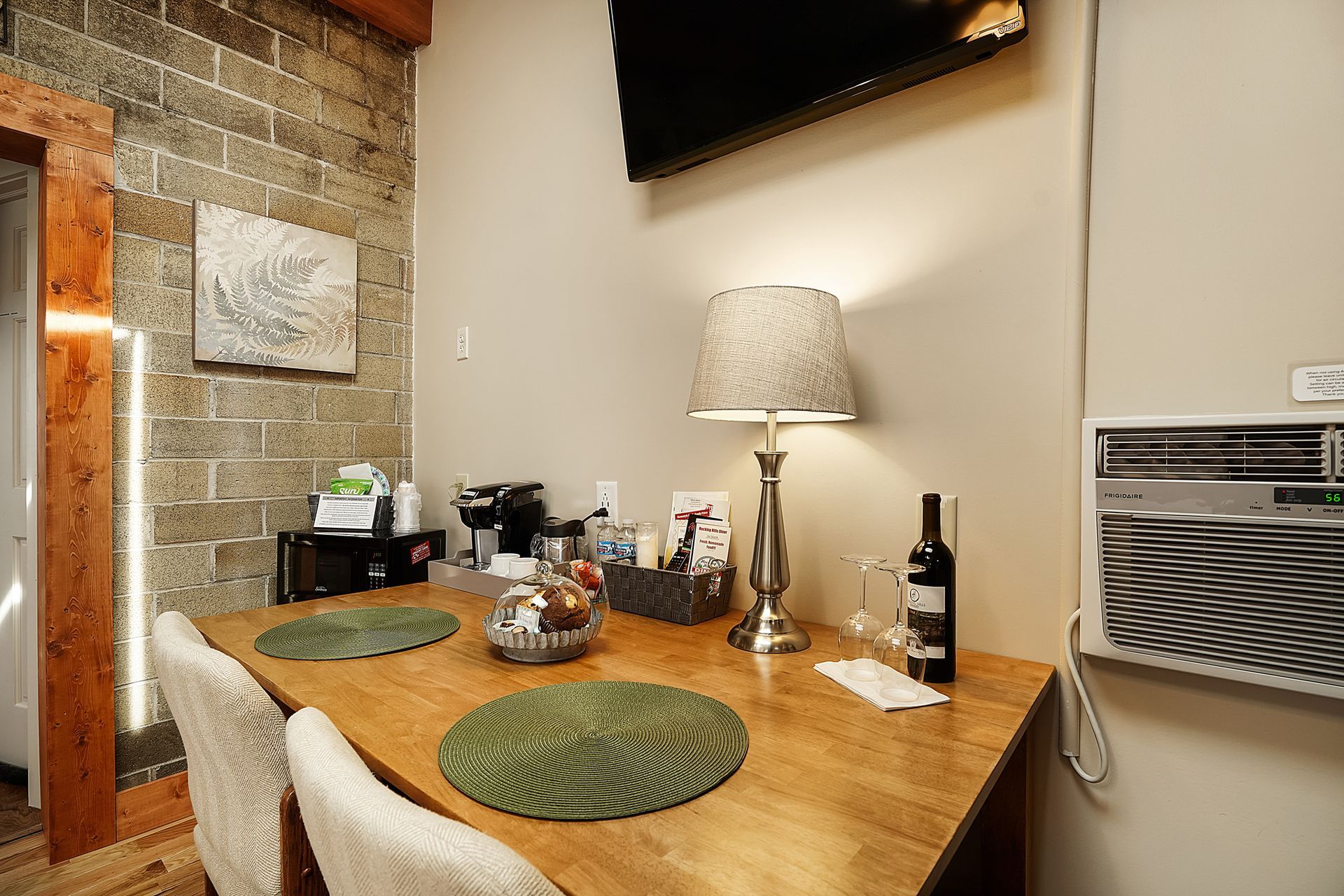 A small dining area with a wooden table, two chairs, and a lamp. A bottle of wine, glasses, and snacks are on the table.