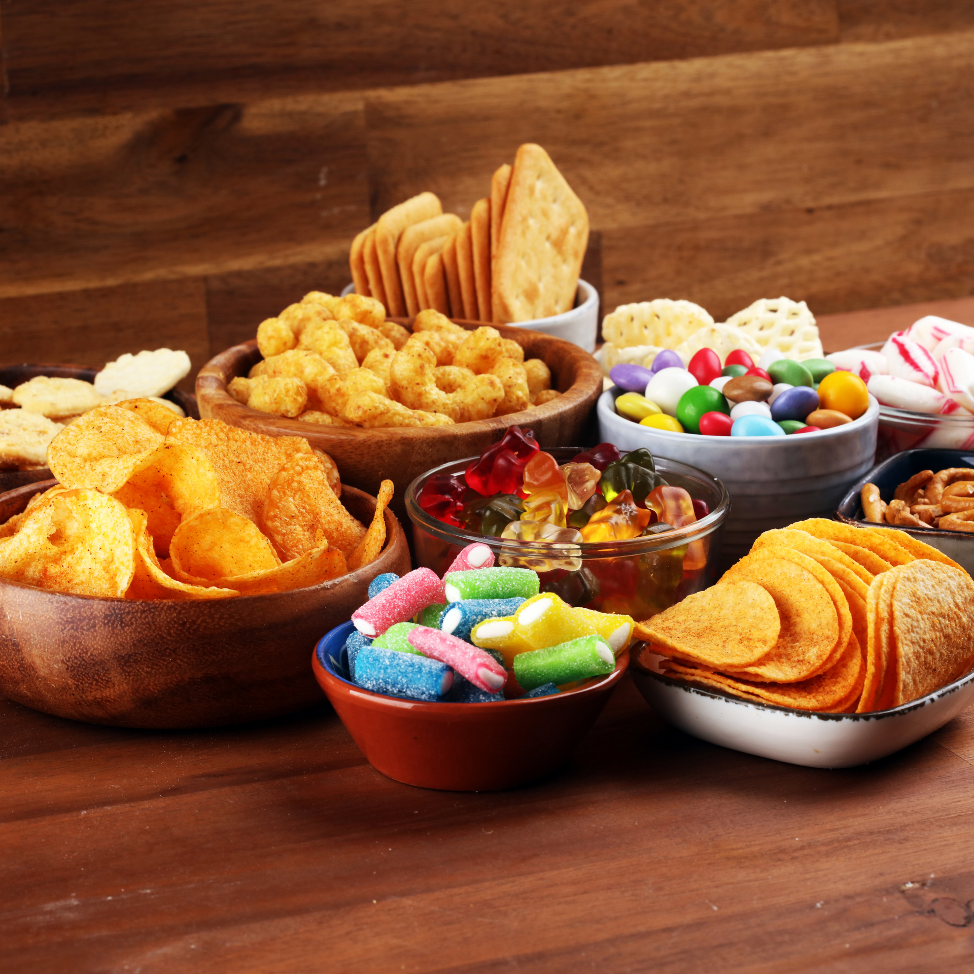 Assortment of snacks in bowls on a wooden surface, including chips, crackers, candies, and nuts.