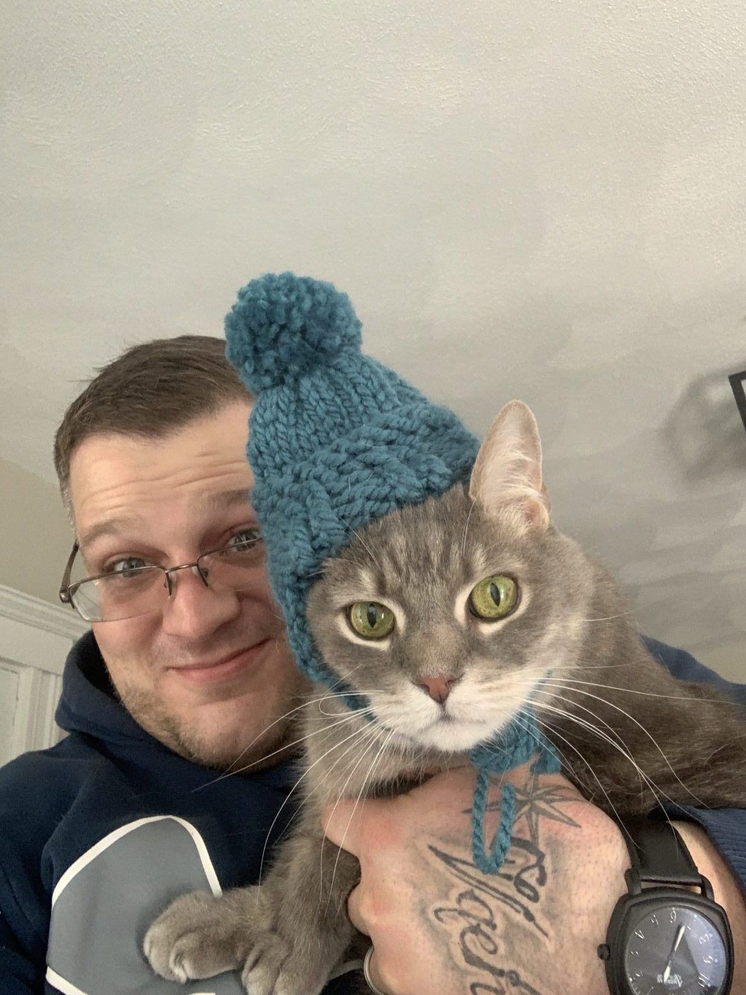 Man in glasses holding a gray cat wearing a blue knitted hat. They are looking at the camera.