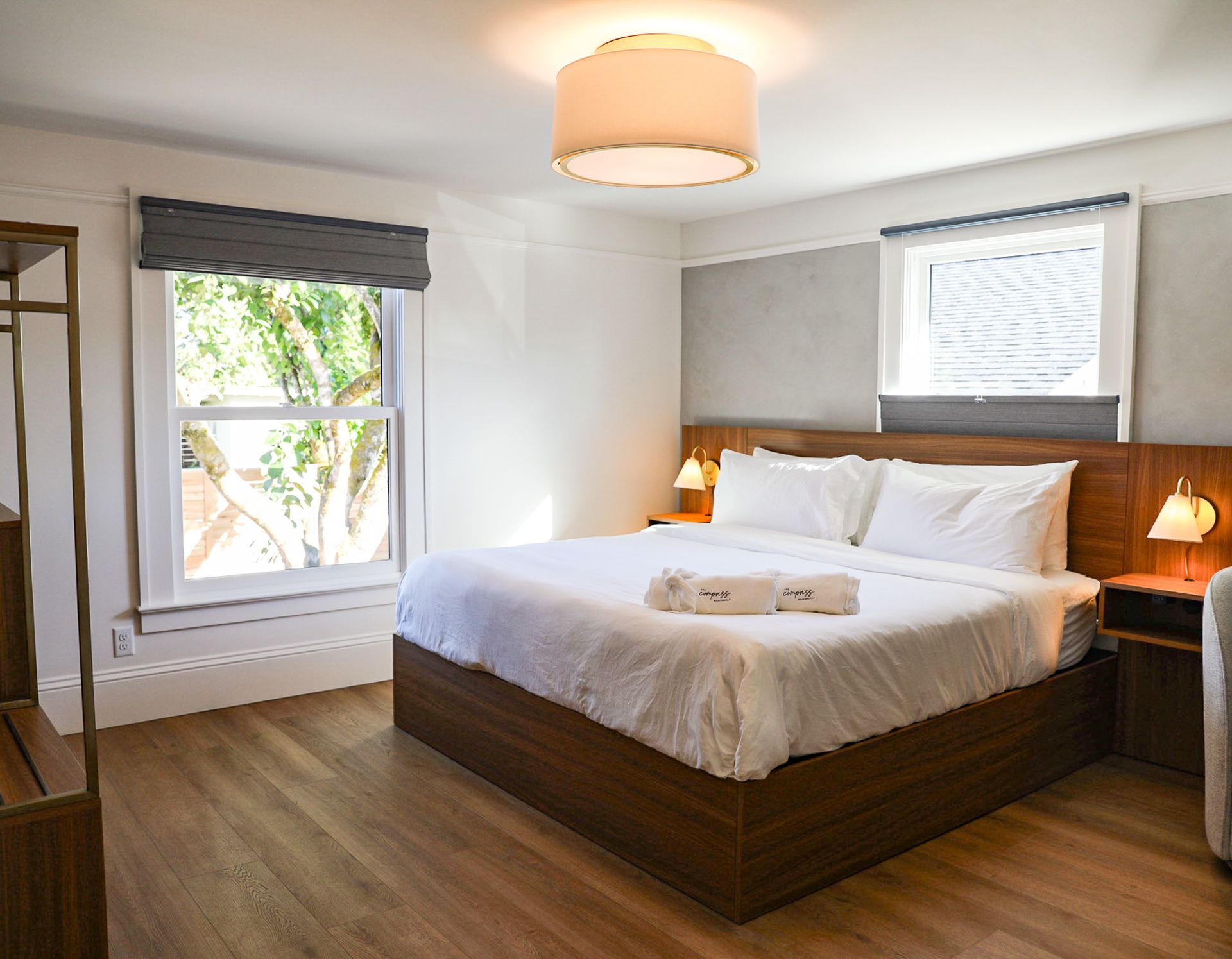 A bedroom with a bed , ottoman , lamps , and two windows.