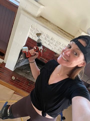 A woman is taking a selfie while holding a drill in front of a fireplace.