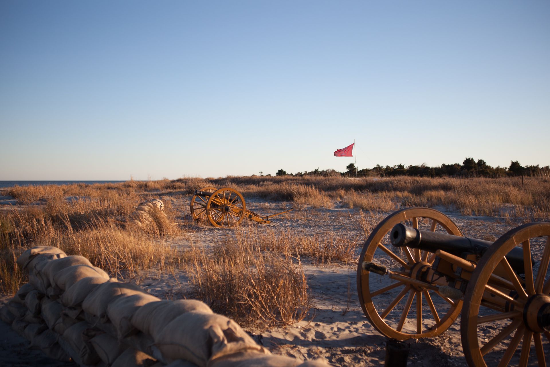 Field with cannons behind sandbags, red flag waving, dry grass, sunny sky.