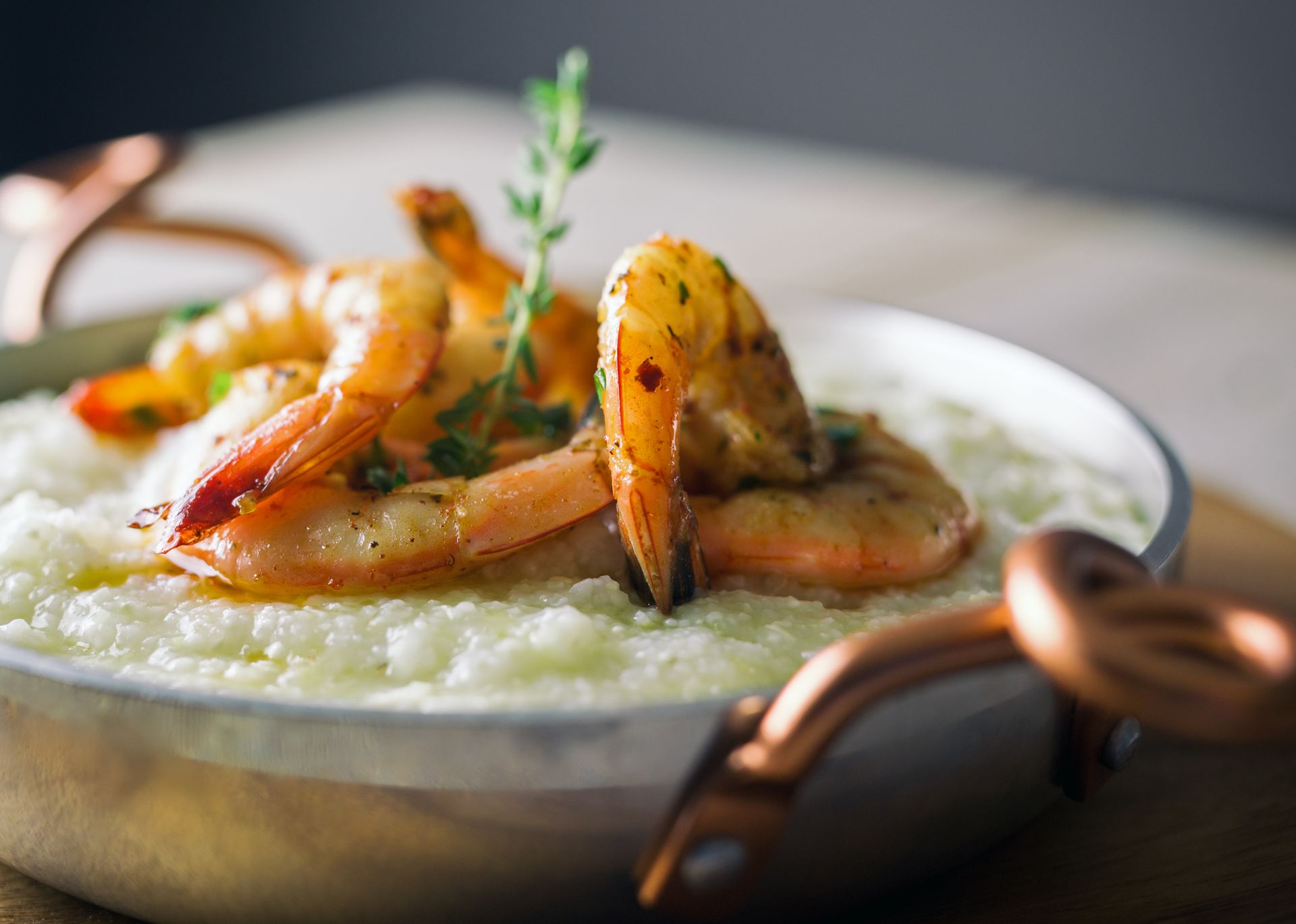 Shrimp and grits in a silver dish with copper handles, garnished with herbs.