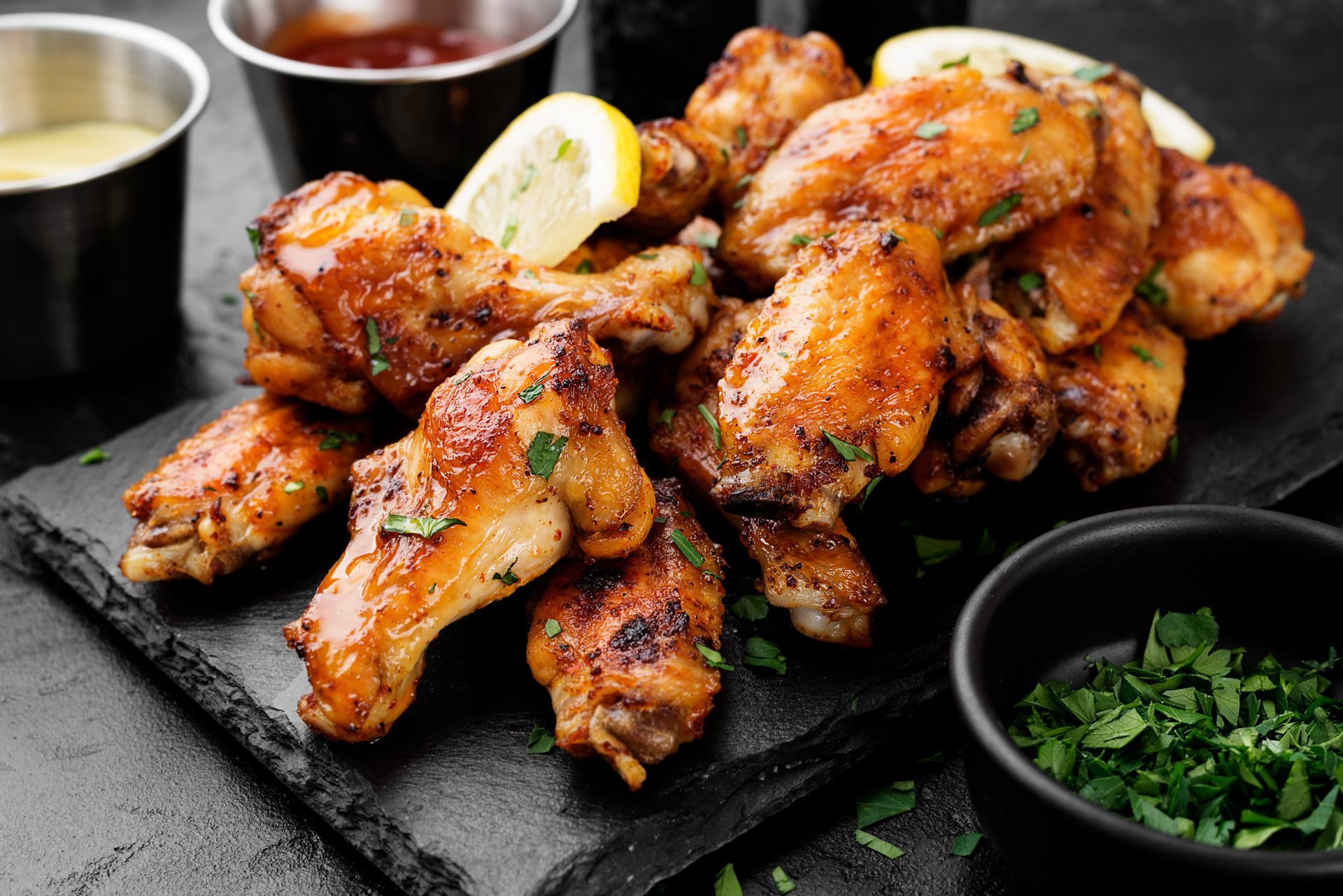 Crispy, golden chicken wings on a black slate, with dipping sauces and lemon slices.