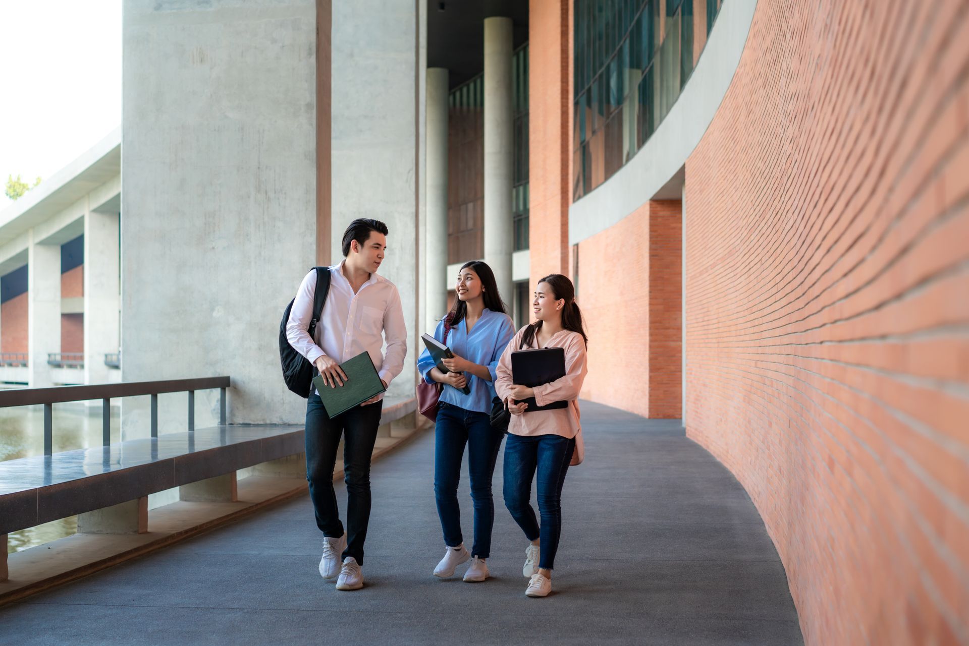Three students walking and talking on a university campus walkway with a brick wall on the right.