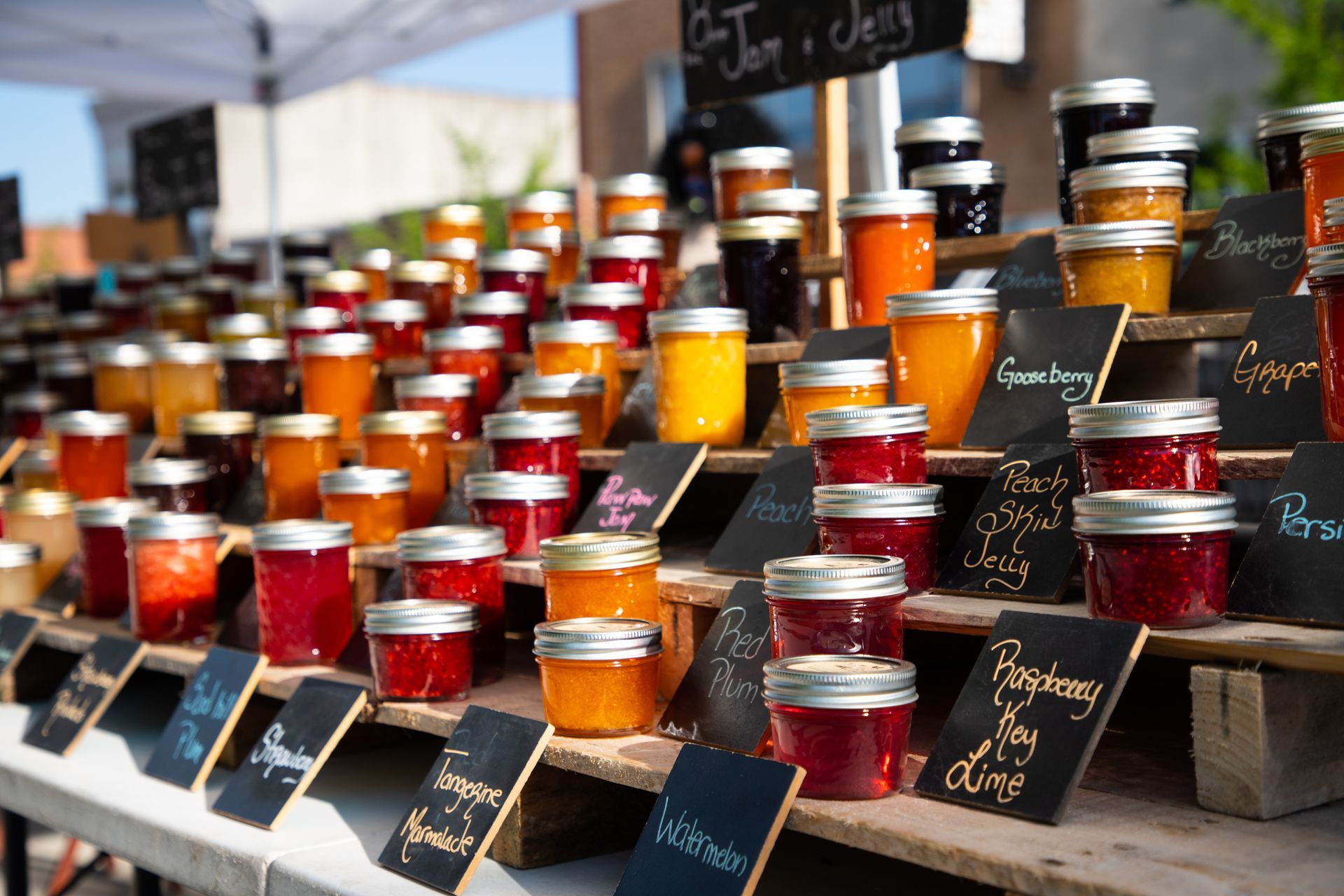 Jars of colorful jams displayed on tiered shelves at a farmers market, with handwritten labels.