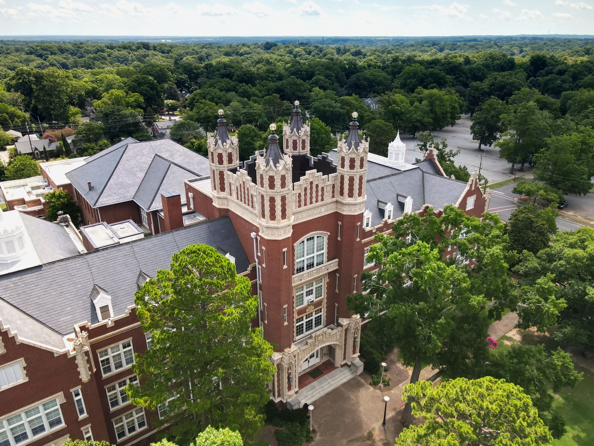 Aerial view of a brick building with a central tower, surrounded by trees and blue sky.