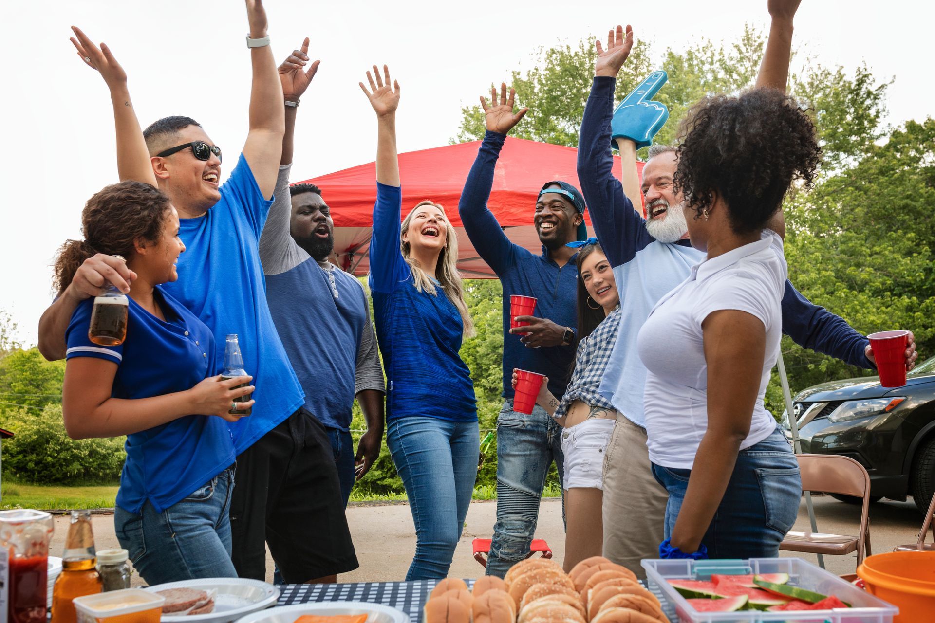 Group of people at a tailgate party, arms raised in celebration; food and drinks on the table.