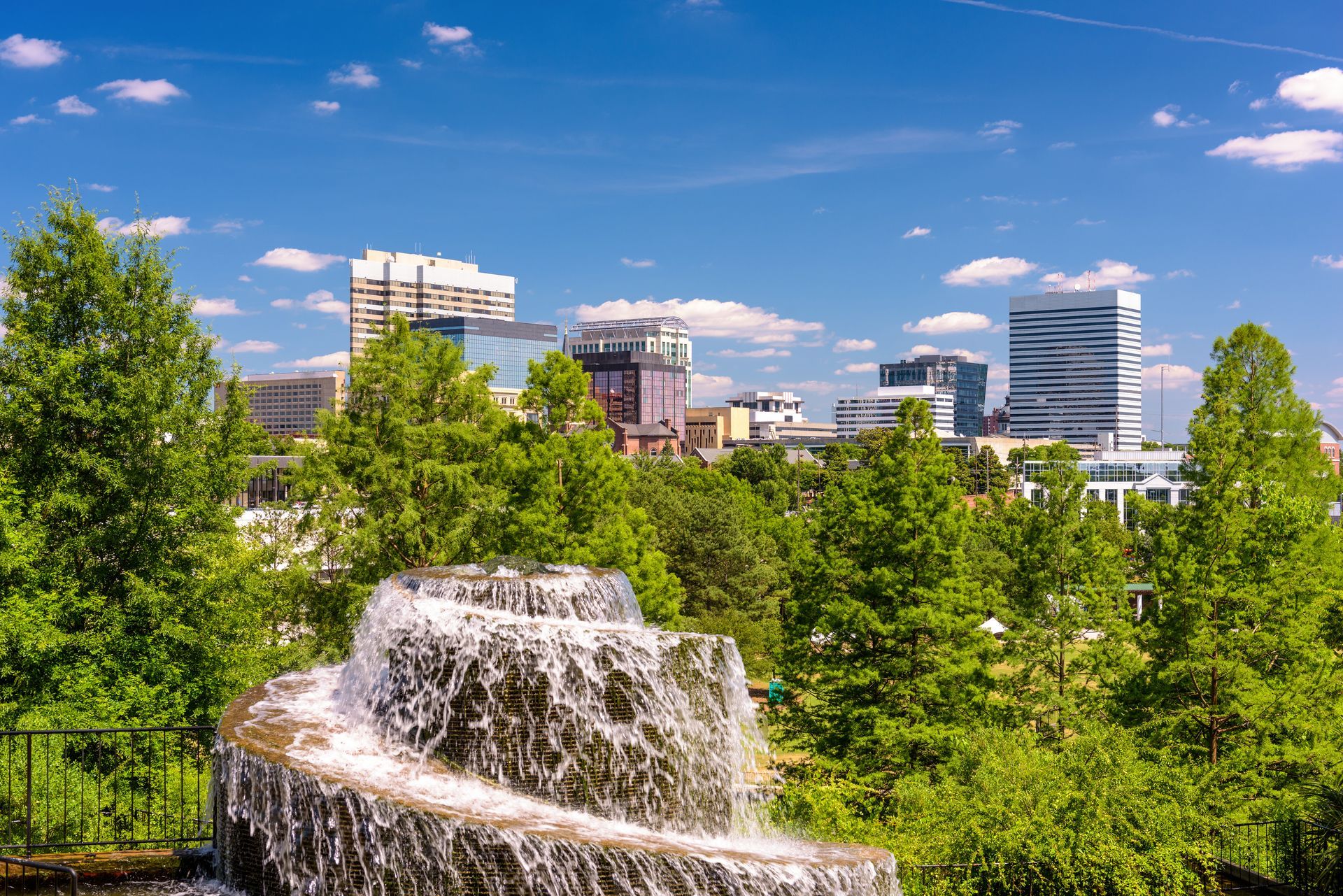 City skyline framed by green trees, water fountain in foreground under blue sky.