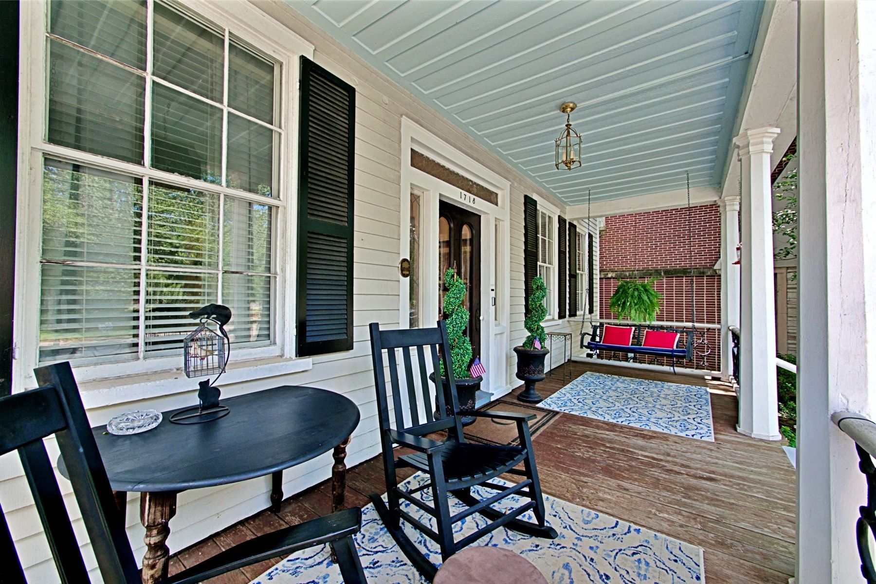 Porch with black rocking chairs, table, and blue ceiling. A swing and potted plants are also visible.