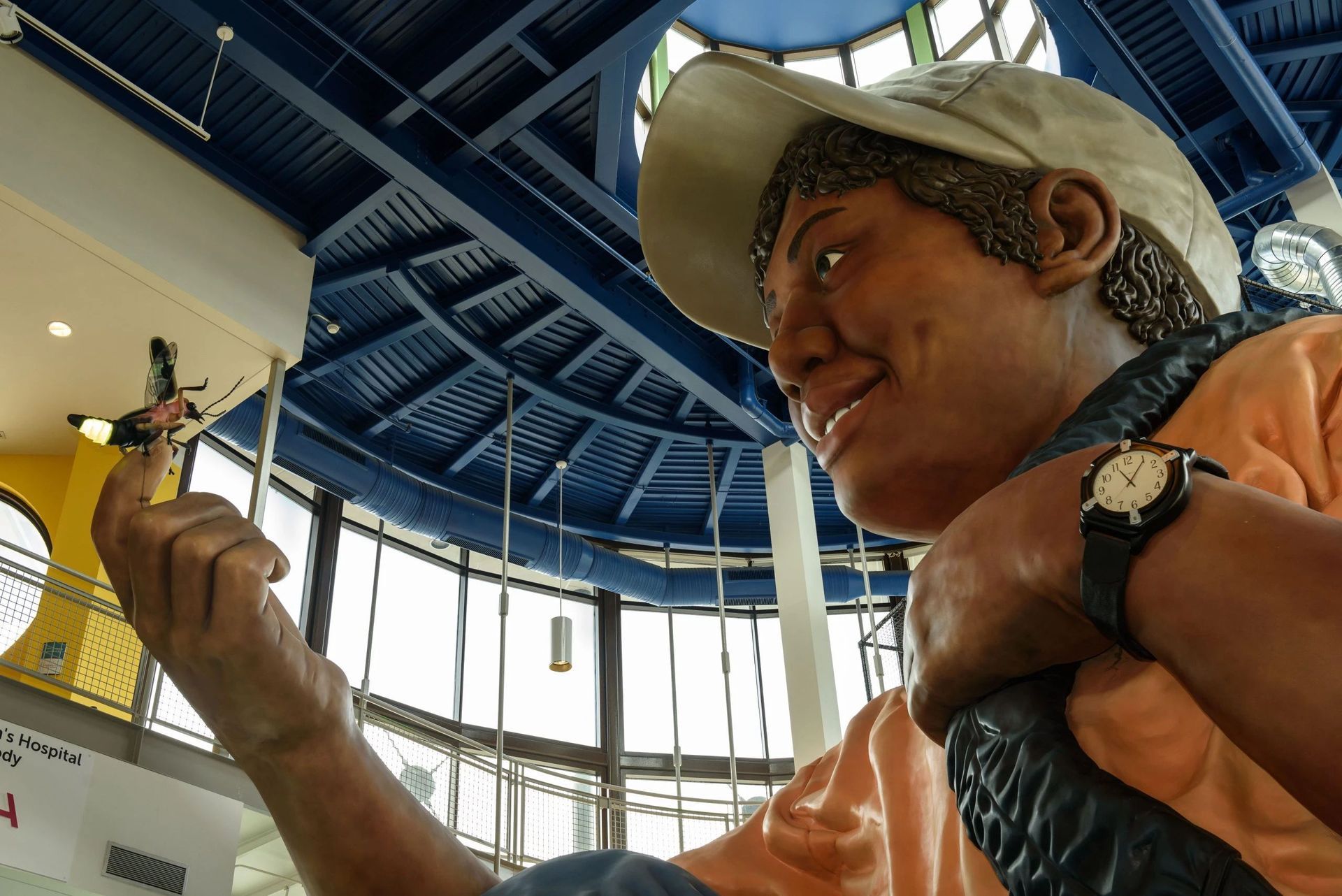 Statue of person smiling at dragonfly, wearing hat, watch, inside building with blue ceiling.