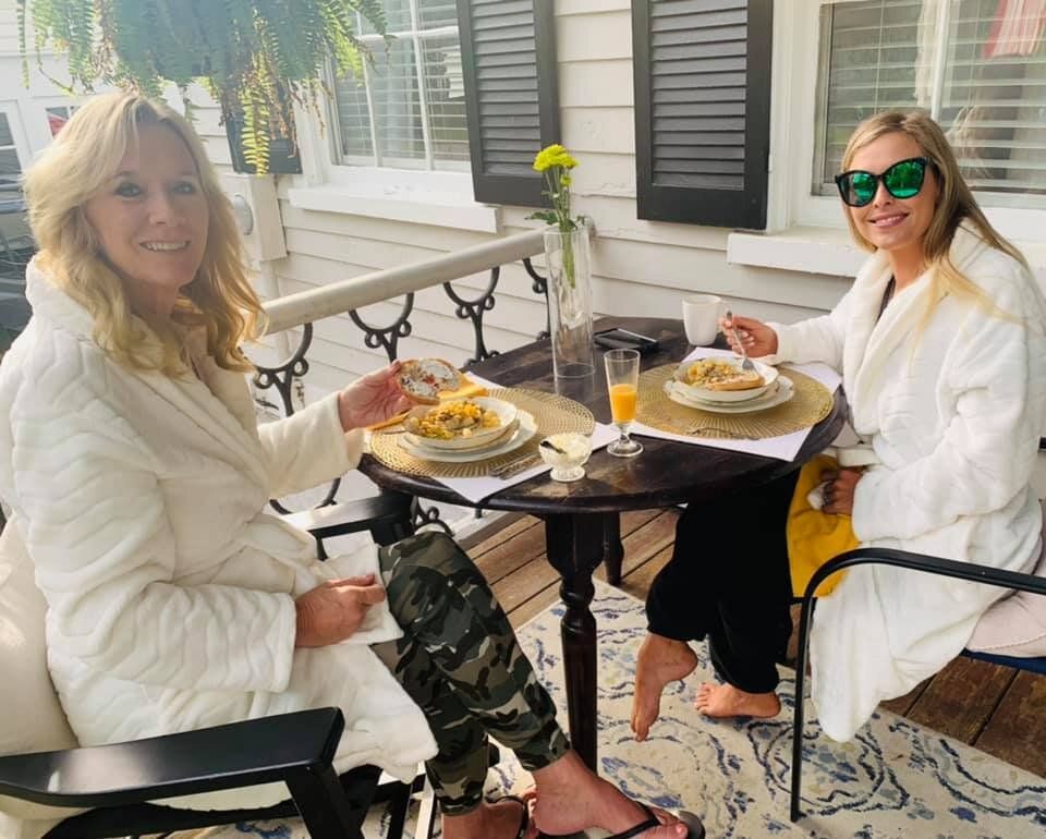 Two women in white robes dine at a small table on a porch. One wears camouflage pants. Sunny setting.