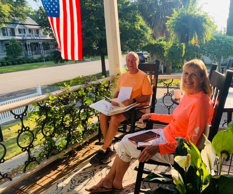 Two people sitting on a porch with a US flag. Man and woman both wearing bright orange, smiling, holding books.